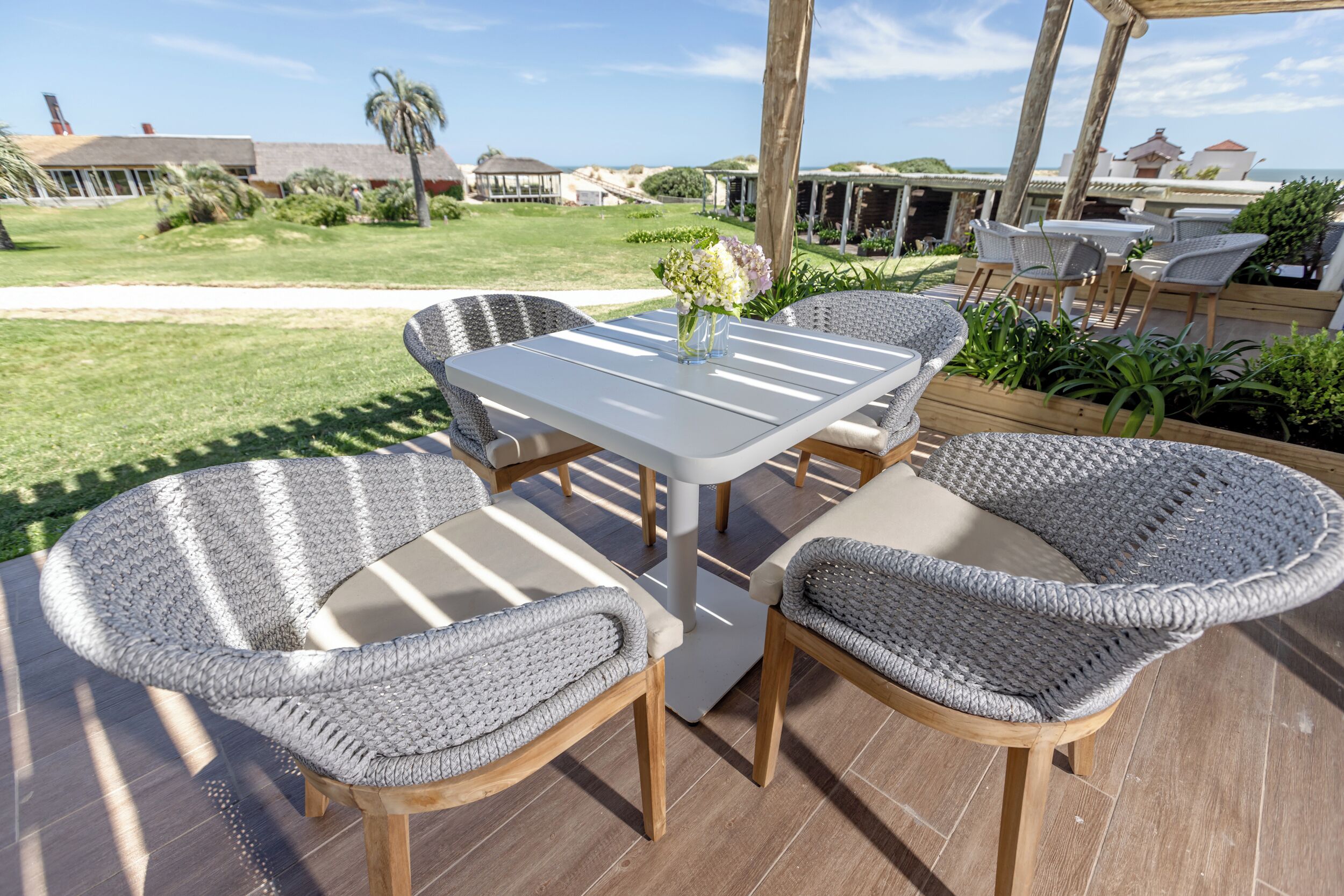 Un patio al aire libre con una mesa blanca y sillas de mimbre, rodeado por una terraza de madera y vista al océano y edificios al fondo.