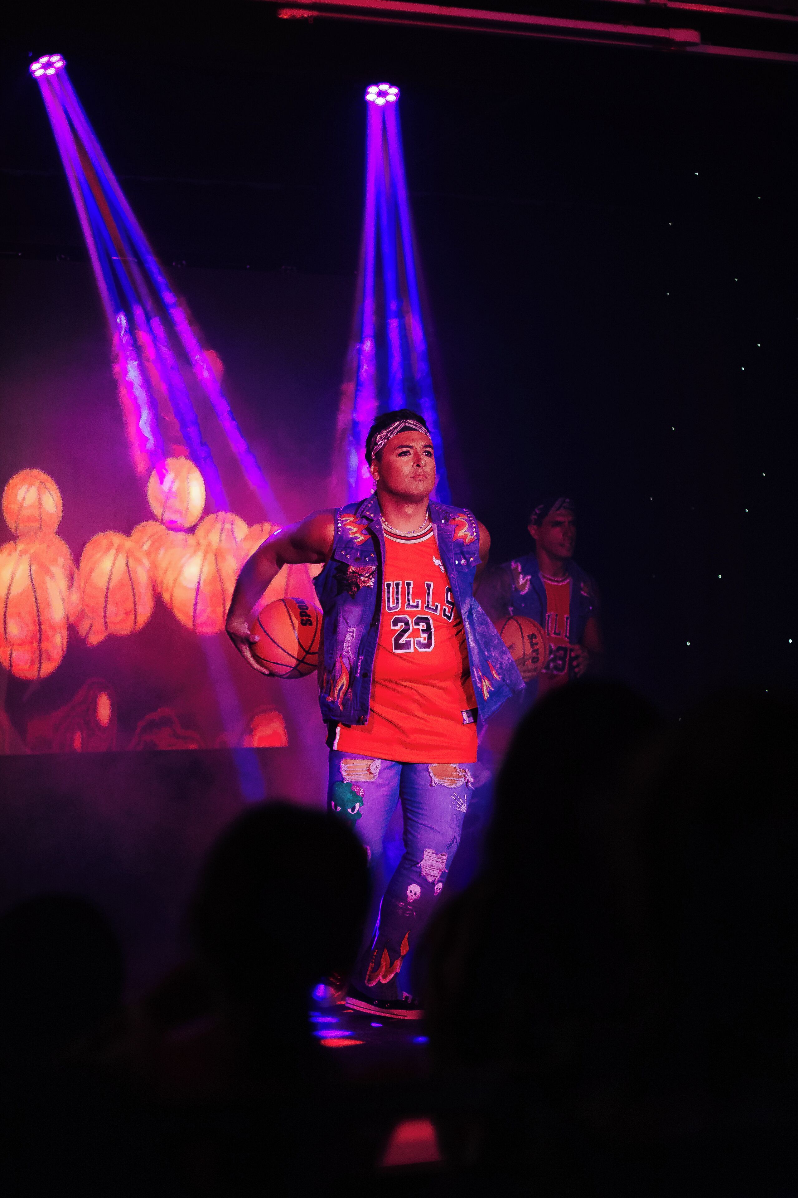 Una persona en el escenario con un balón de baloncesto, vestida con una camiseta roja con el número 23 y un chaleco, en un ambiente tenuemente iluminado con luces moradas.