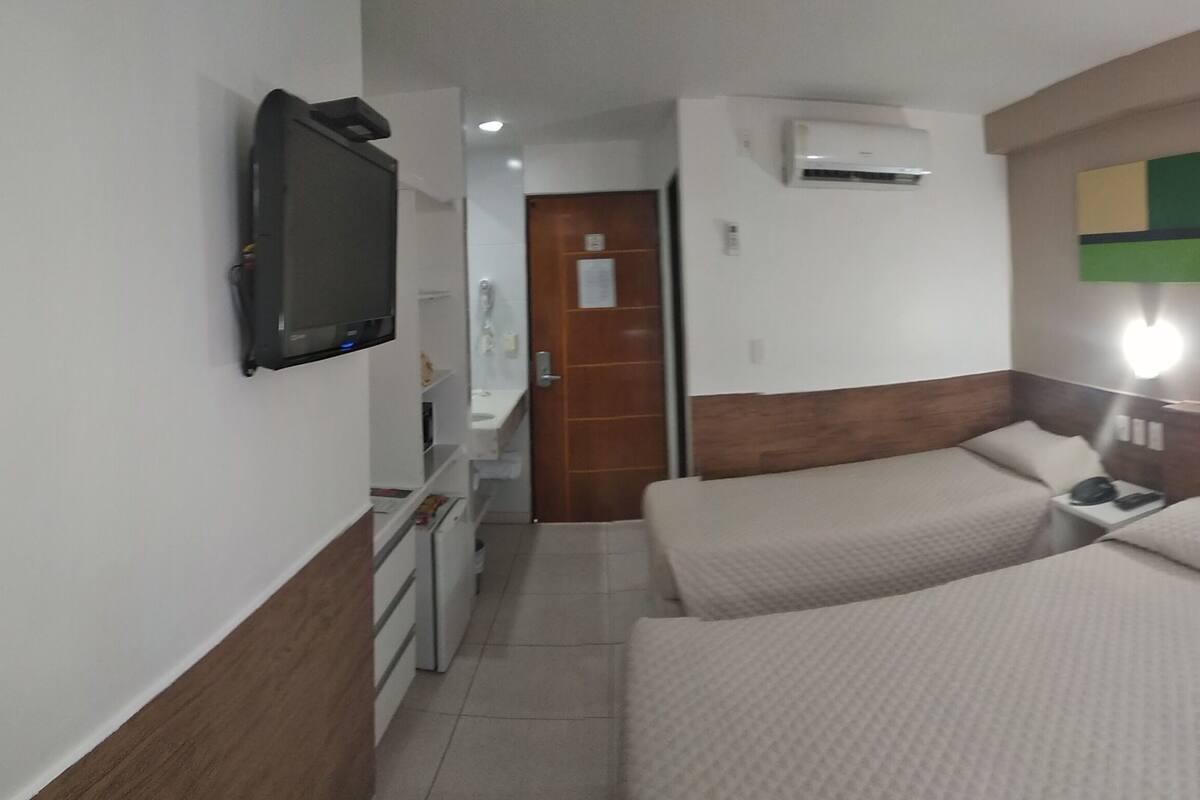 A hotel room with two beds, a TV, an air conditioner, a bedside table with a phone, and a wall-mounted light.