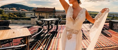 A person in a white dress standing on a rooftop terrace with a colorful striped carpet, tables, and chairs.