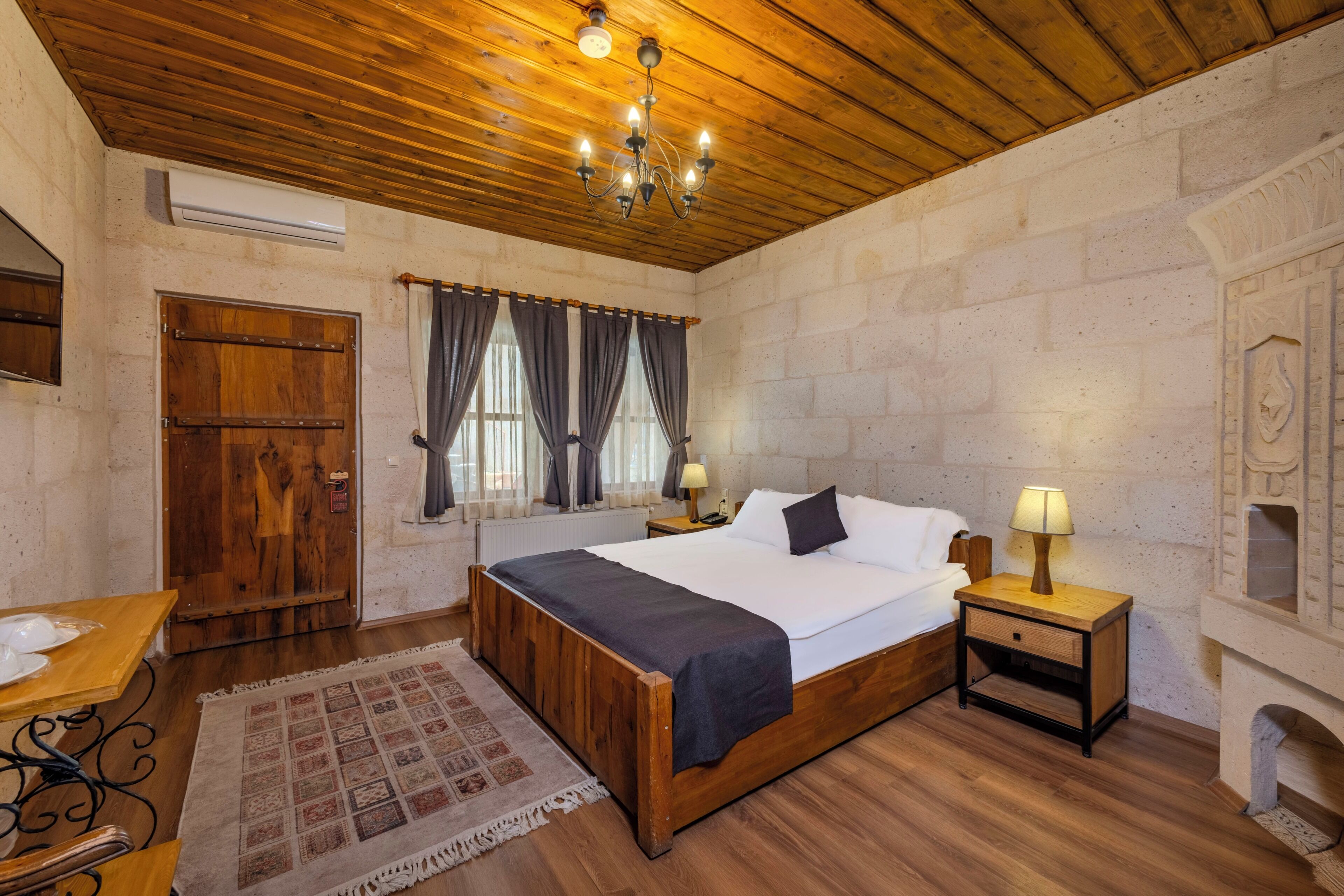 A bedroom with a wooden ceiling, a bed with white linens, two bedside lamps, a wooden nightstand, and a patterned rug.
