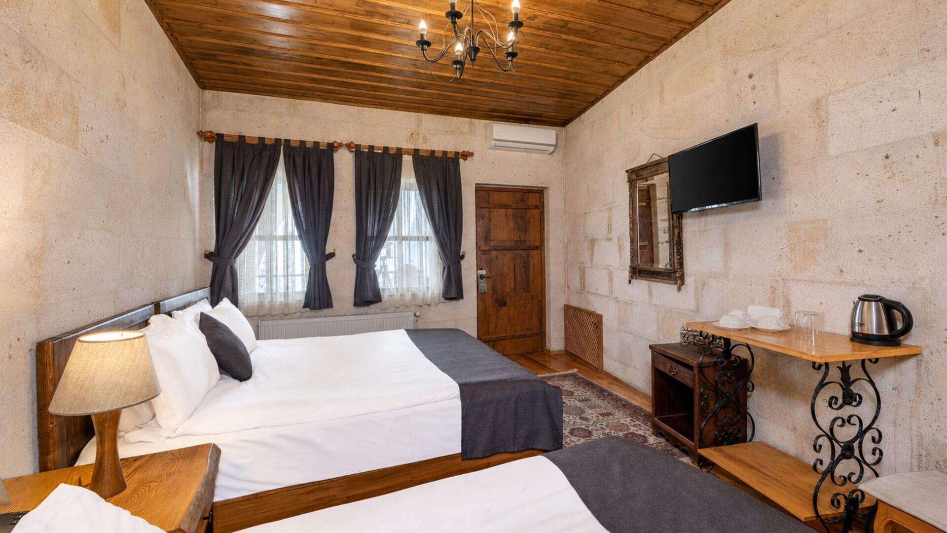 Quarto de hotel com duas camas, teto de madeira, televisão, uma mesa pequena com chaleira e janela com cortinas.