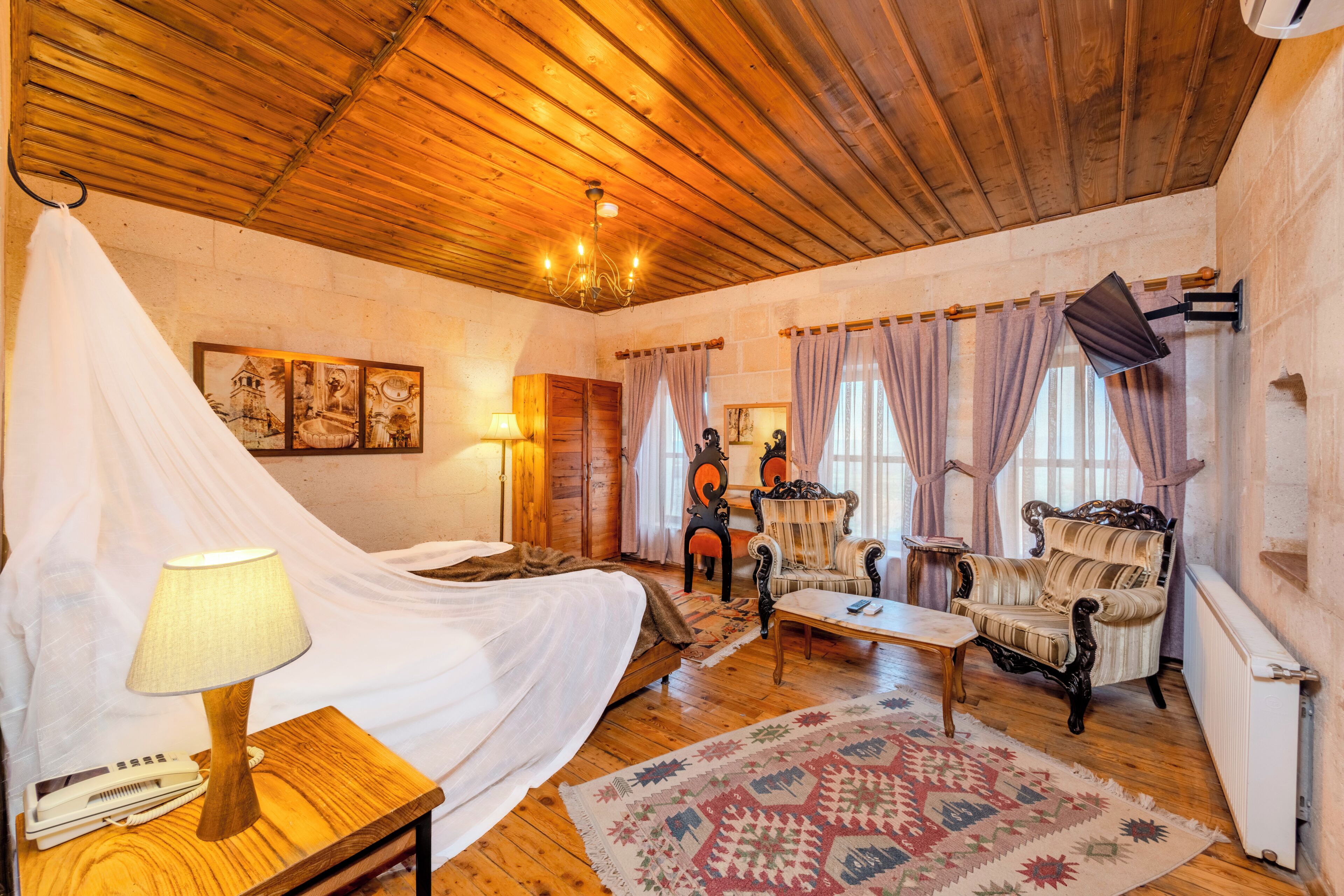 A room with a wooden ceiling, a bed with a white canopy, a table with a lamp, two chairs, and a patterned rug.