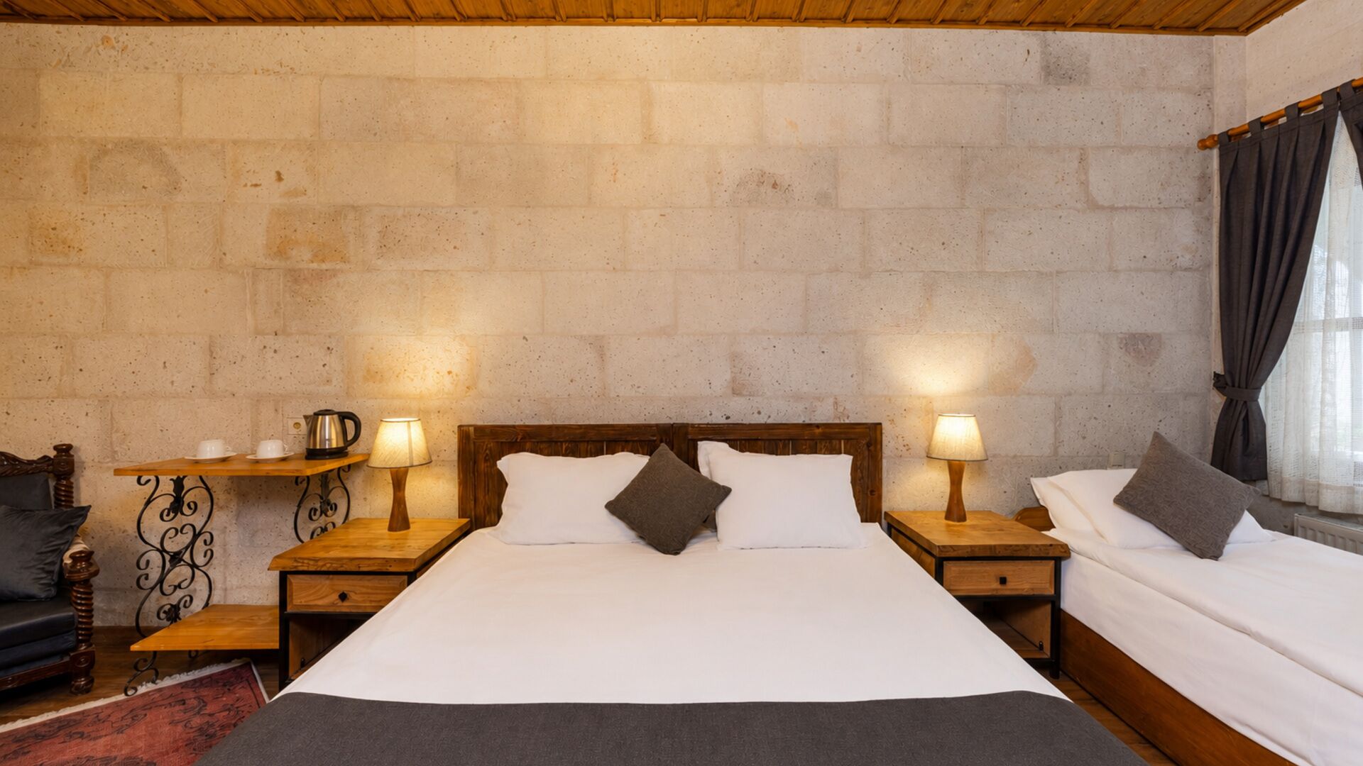 A double room with a stone wall, wooden headboard, two bedside tables with lamps, a small shelf, and a black leather sofa.