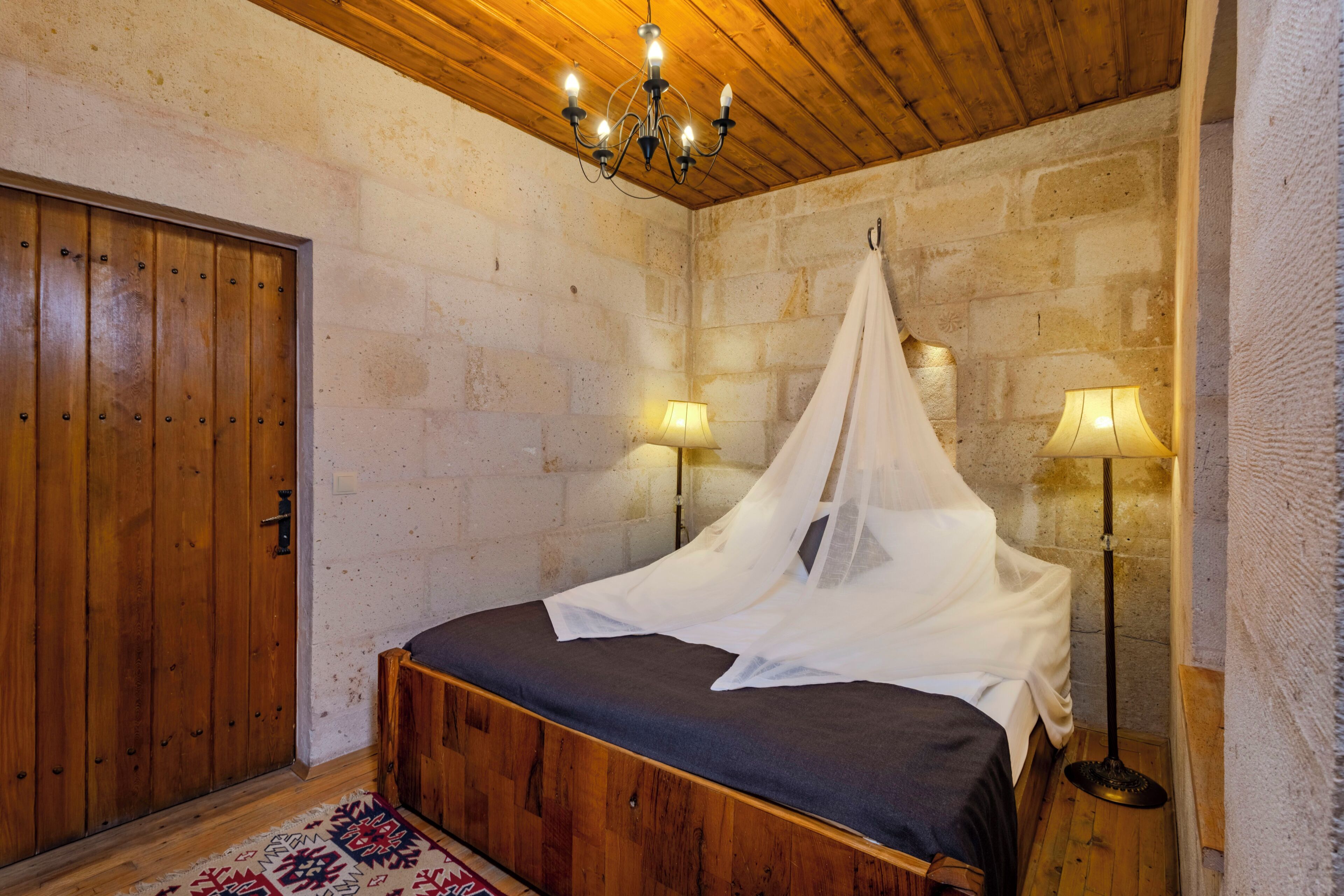 A bedroom with a wooden bed, a canopy, a nightstand with a lamp, and a stone wall.
