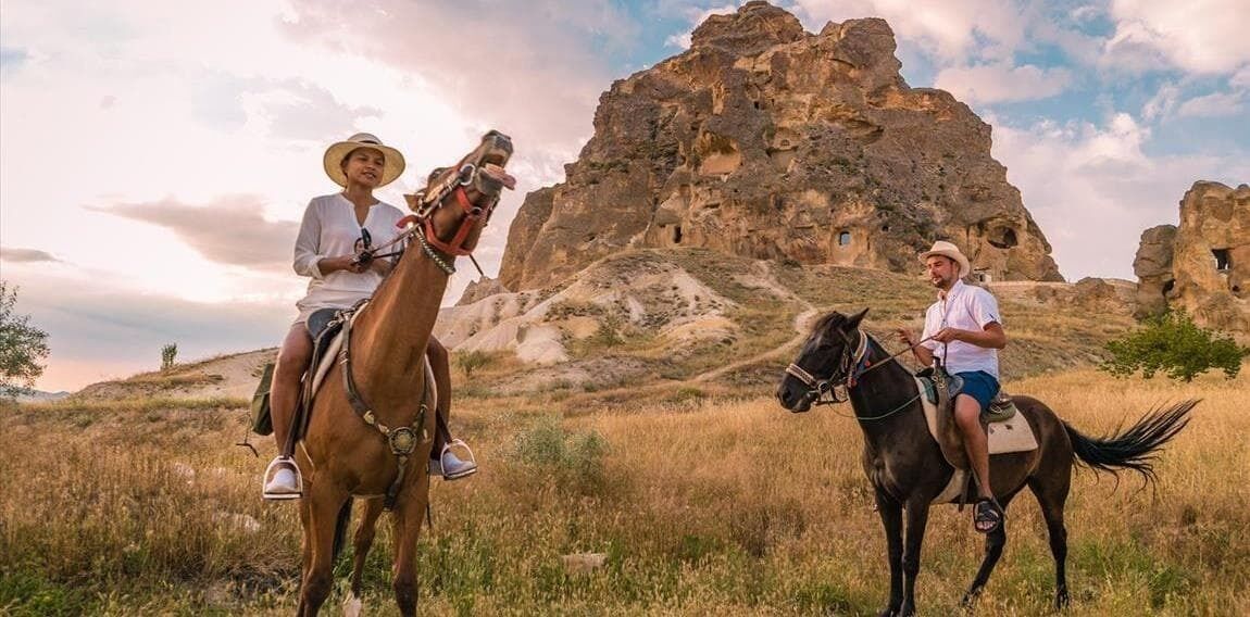 Two people riding horses in a natural landscape with rocky formations in the background.