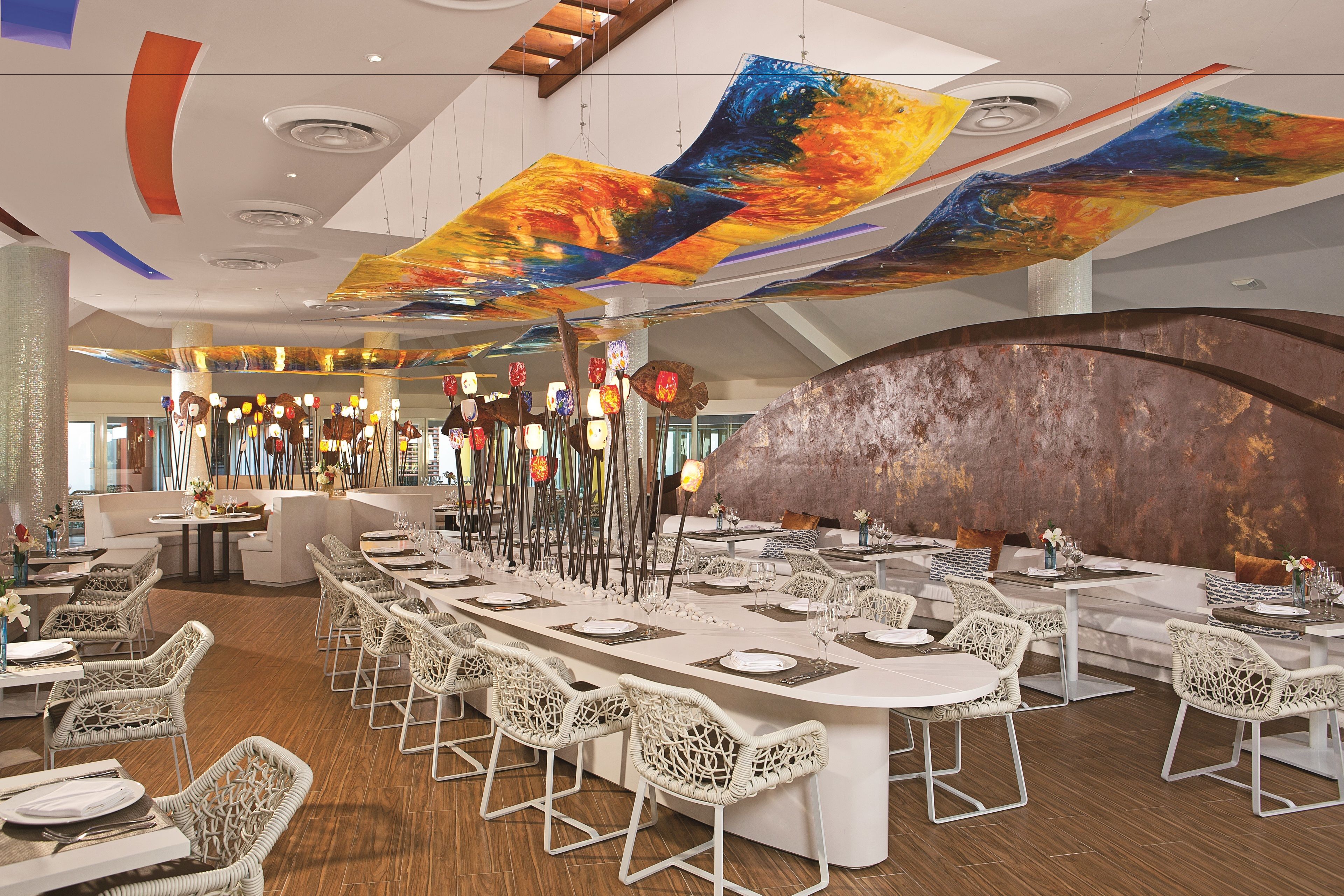 A modern dining area with a long table set for multiple guests, surrounded by wicker chairs, and a large abstract painting hanging from the ceiling.