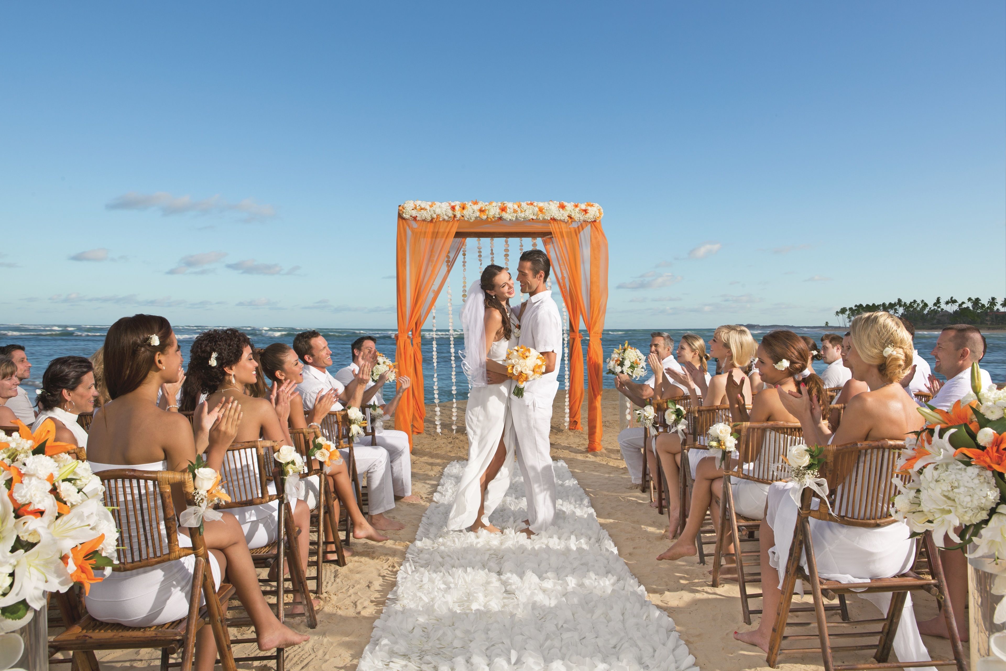 Une cérémonie de mariage sur la plage, avec une allée blanche et des invités assis sur des chaises en bois.