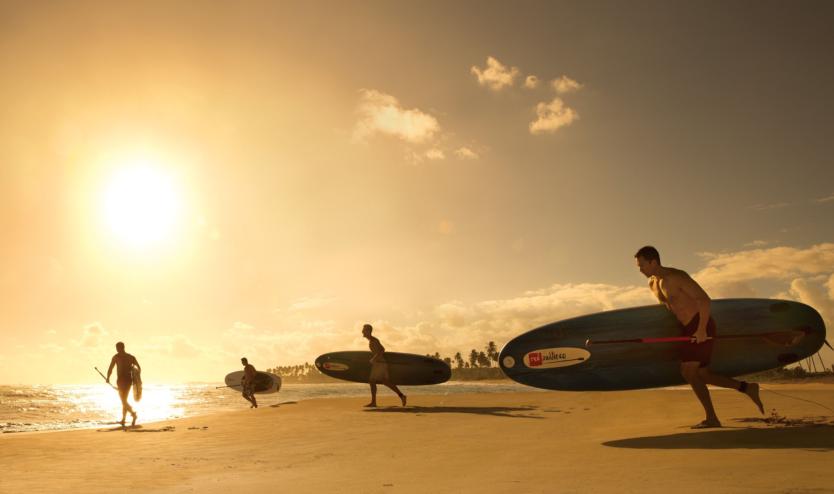 Drie mensen wandelen over een strand met surfplanken, een heldere lucht en een ondergaande zon.
