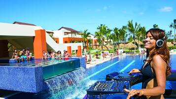 Relax by the pool surrounded by palm trees and thatched umbrellas, with a DJ setup for entertainment.