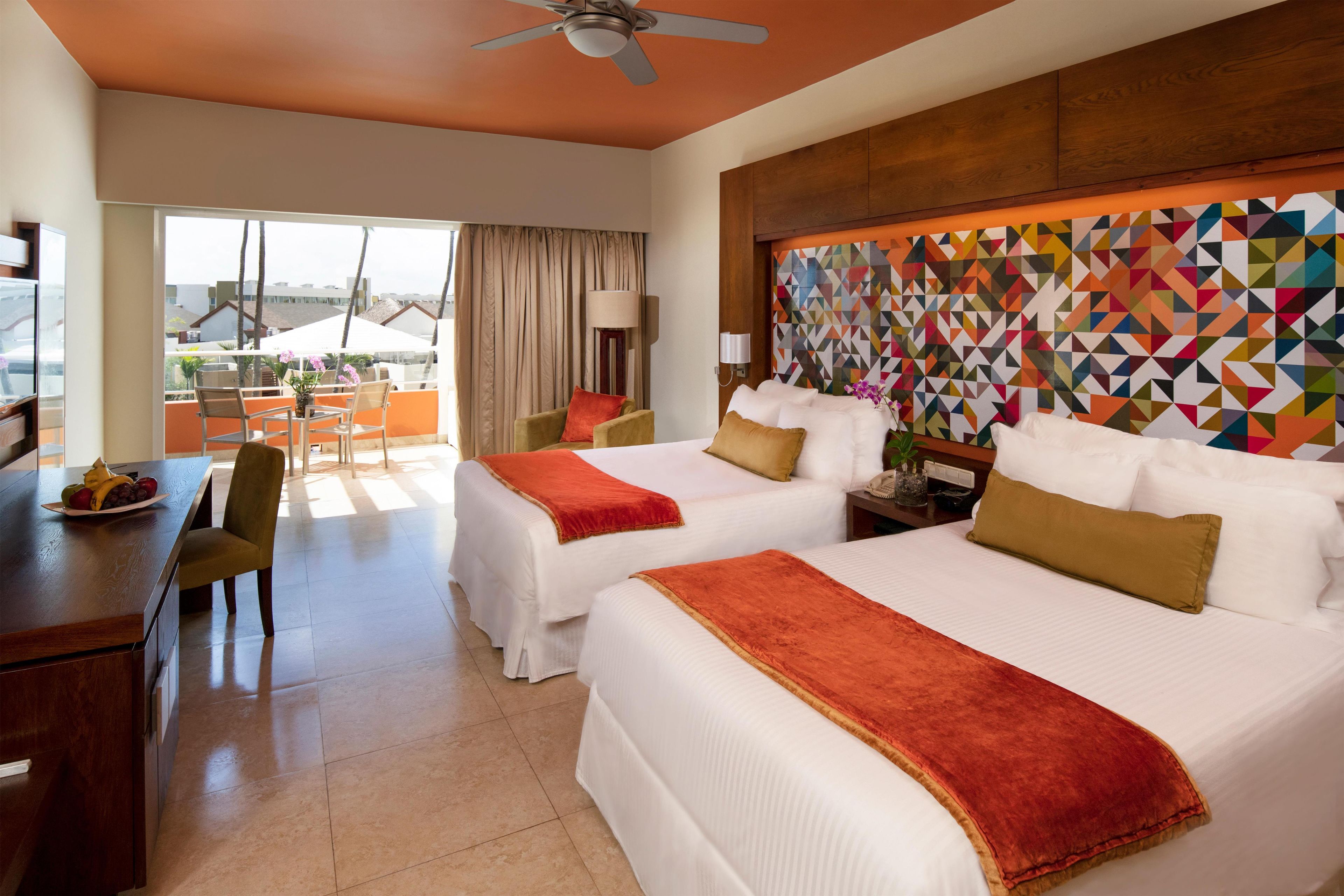 A hotel room with two beds, a desk, chairs, a balcony with a view, and a colorful geometric wall.