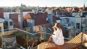 A person sitting on a wooden deck with a glass balustrade overlooking a cityscape with red-roofed buildings and a distant church spire. - The Emblem Prague Hotel  (Prague)