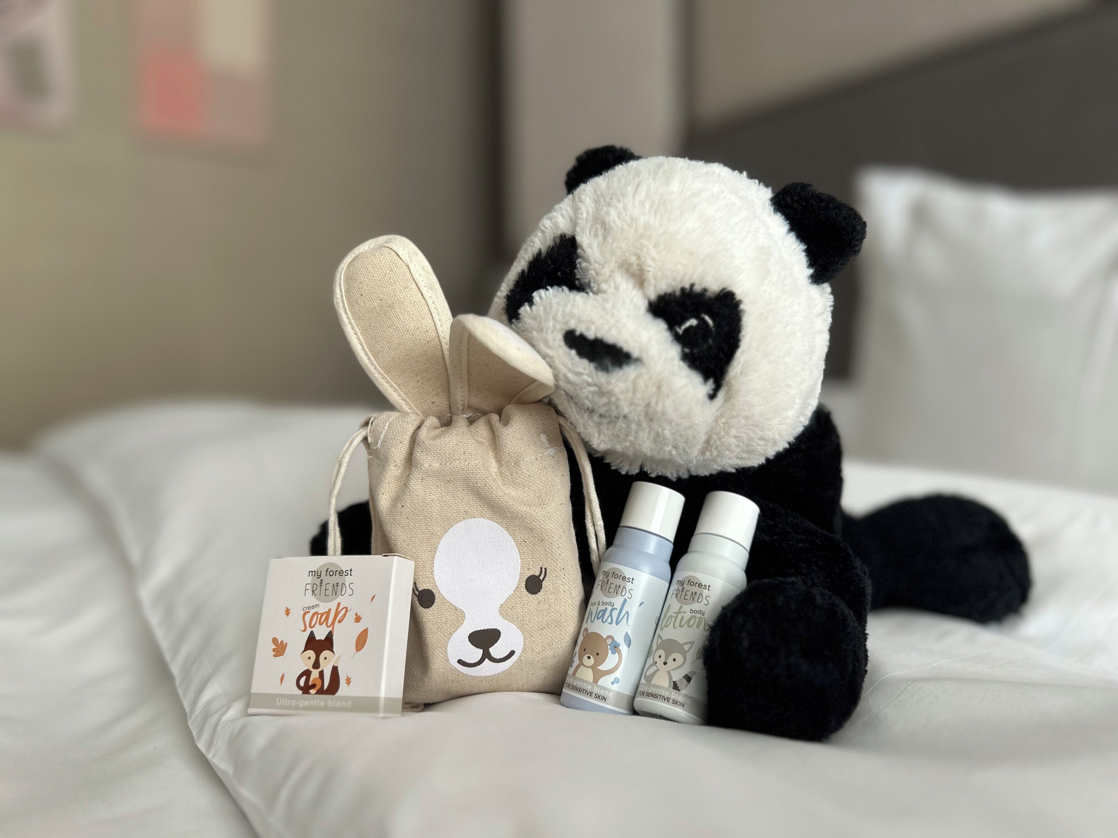 Un peluche de panda sobre una cama con una pequeña bolsa y dos botellas de jabón.