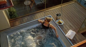 A person relaxing in a hot tub on a wooden deck with a glass wall, a table, and a bottle of champagne. - The Emblem Prague Hotel  (Prague)