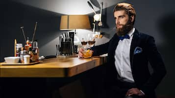 A man in a dark suit and bow tie sitting at a wooden bar with a lamp, glasses, and bottles.