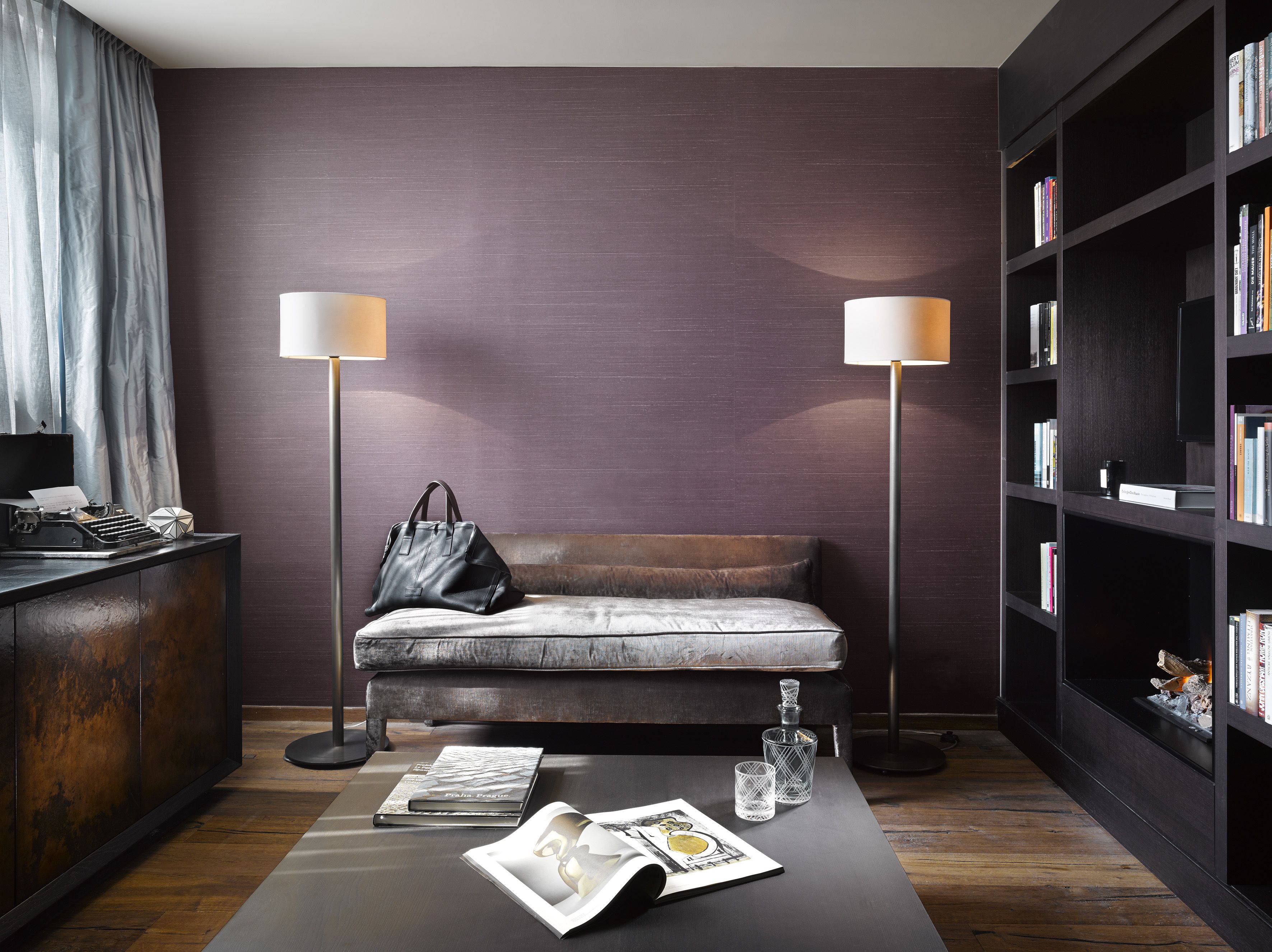A modern reading area with a sofa, two floor lamps, a bookshelf, and a small table with a magazine and glasses.