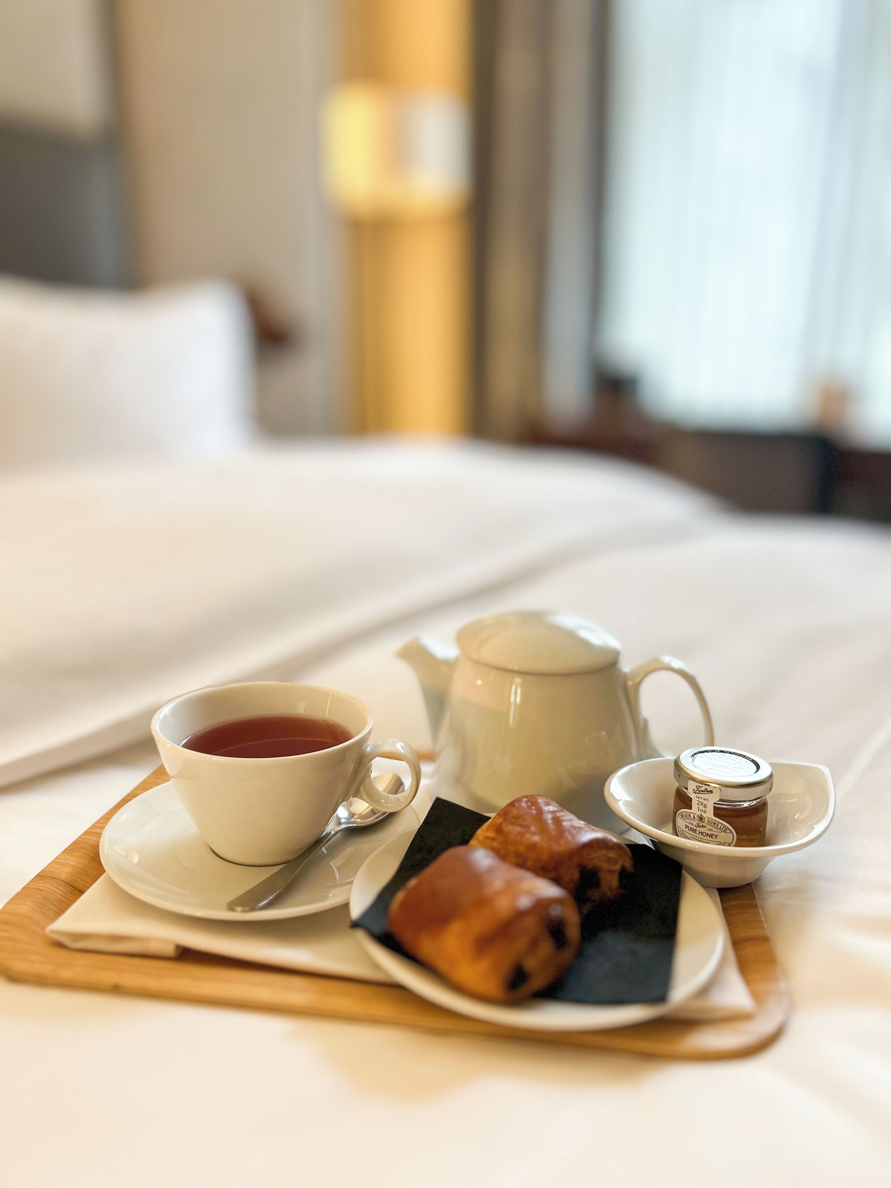 A neatly arranged breakfast tray on a bed with a cup of tea, a teapot, a small jar of honey, and a pastry.
