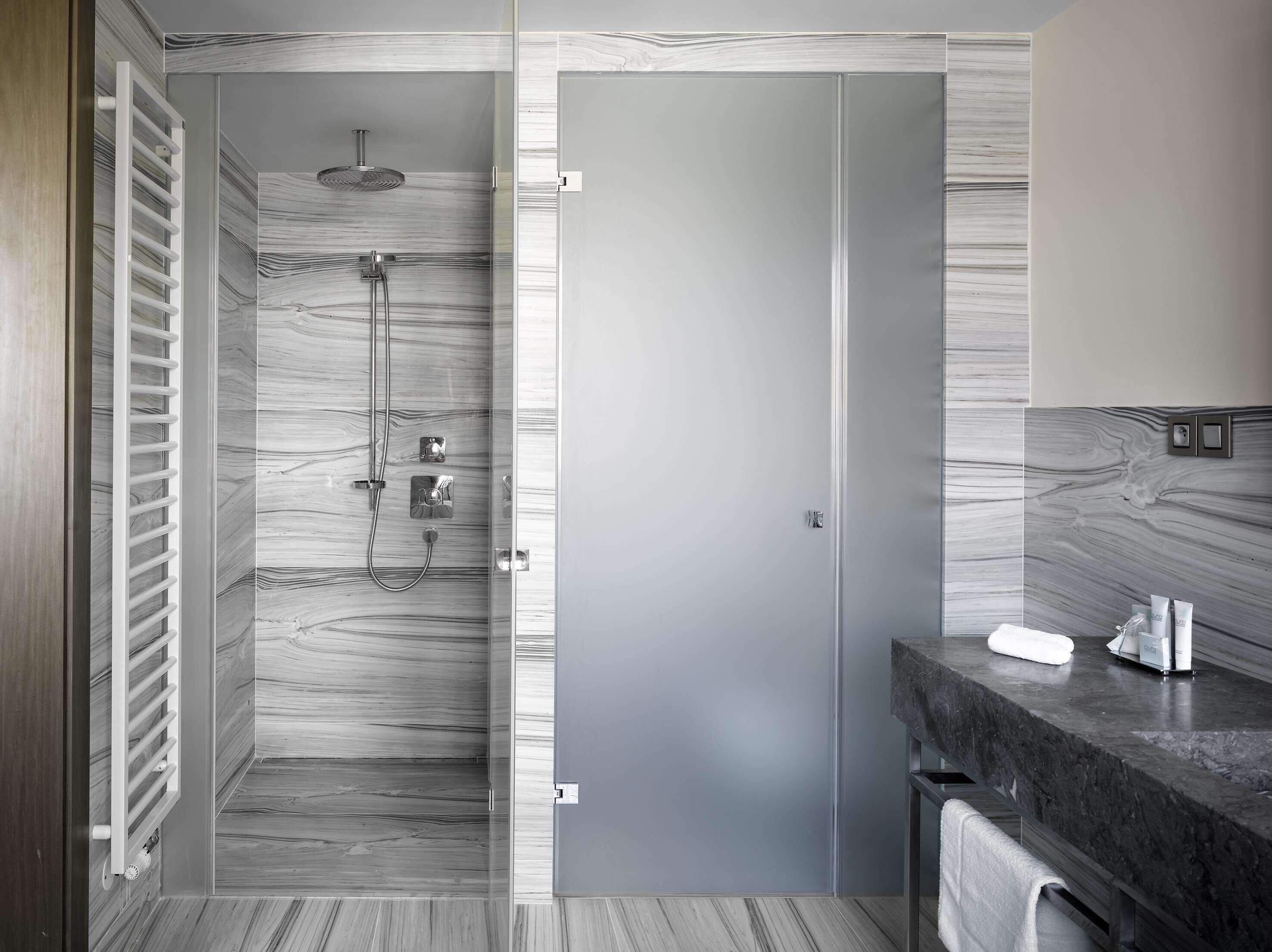 Une salle de bain moderne dotée d’une cabine de douche en verre, d’un espace de douche carrelé en marbre, d’un plan de travail en granit foncé et d’un porte-serviettes.