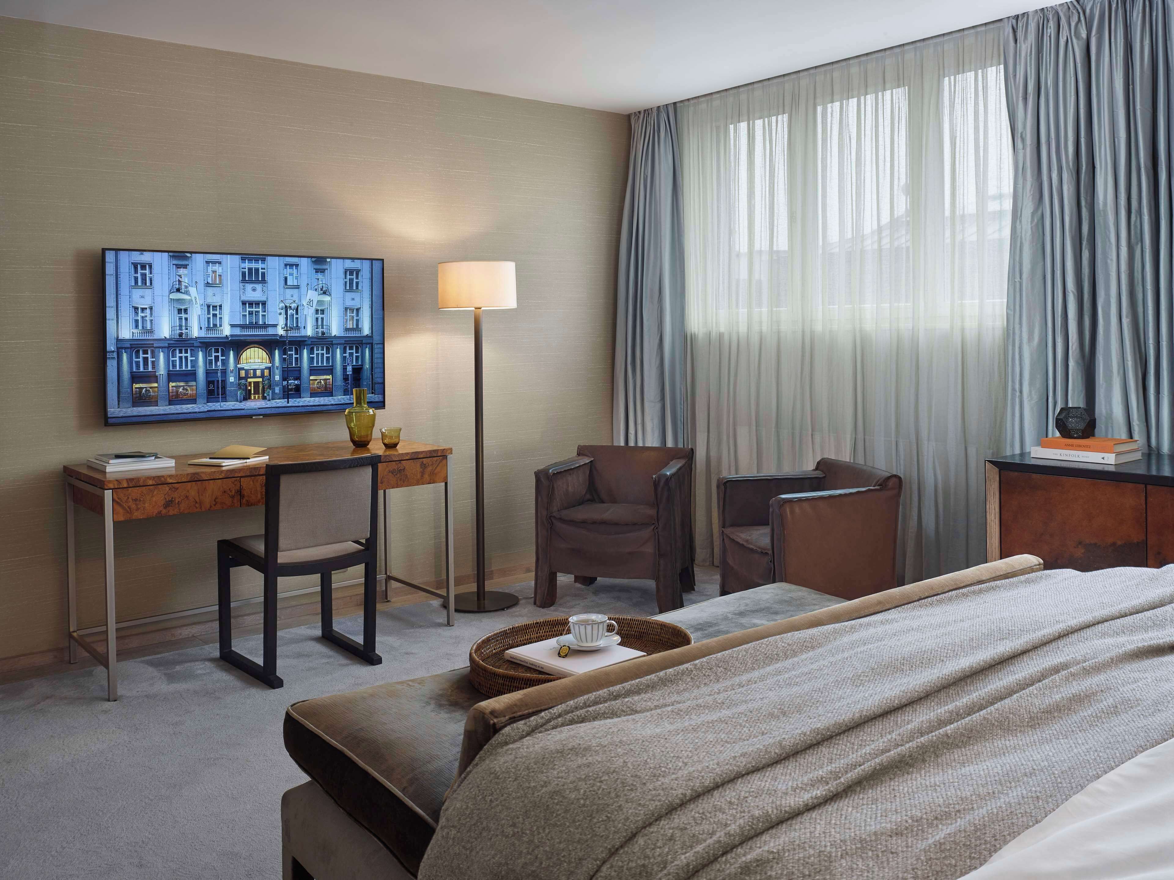 A hotel room with a bed, a desk with a chair, a lamp, a painting on the wall, and a window with curtains.