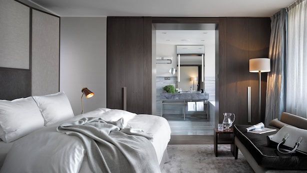 A modern hotel room with a large bed, a desk, a desk lamp, a nightstand, a mirror, and a bathroom visible through an open door. - The Emblem Prague Hotel  (Prague)