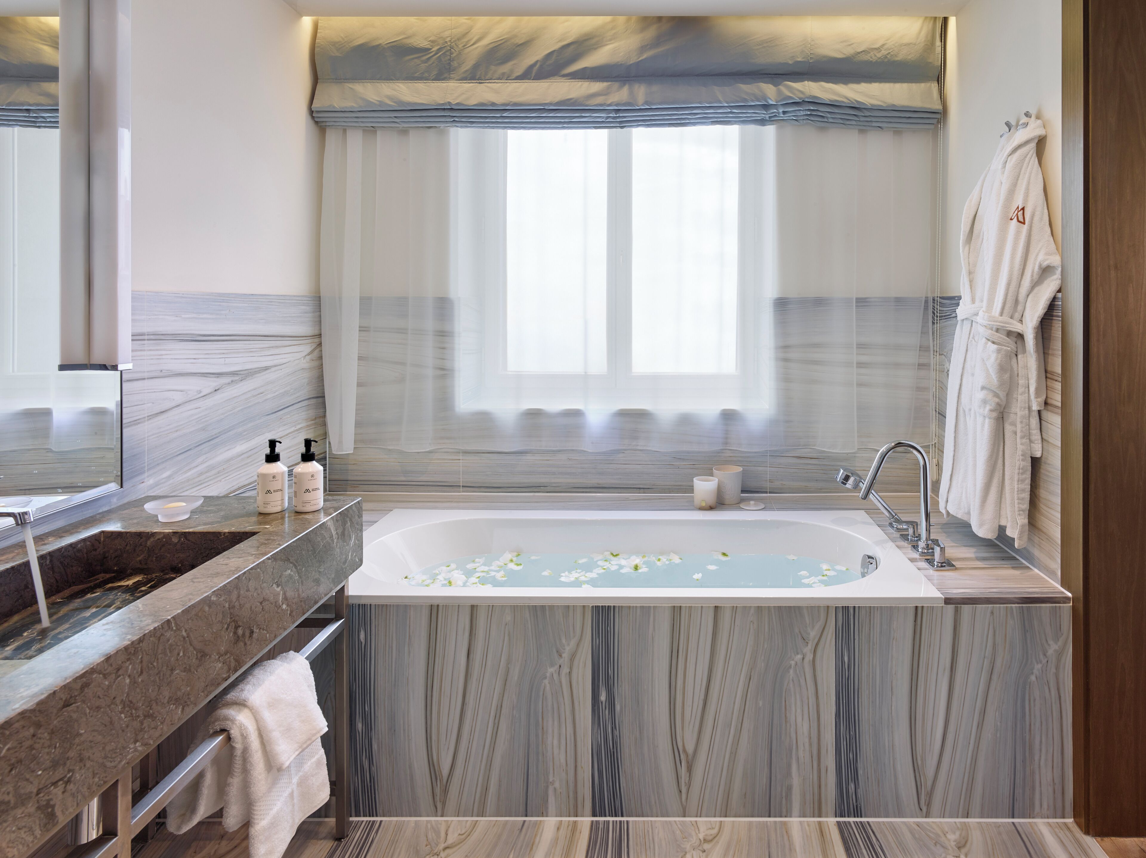 A modern bathroom with a marble vanity, a freestanding bathtub, and a large window with sheer curtains.