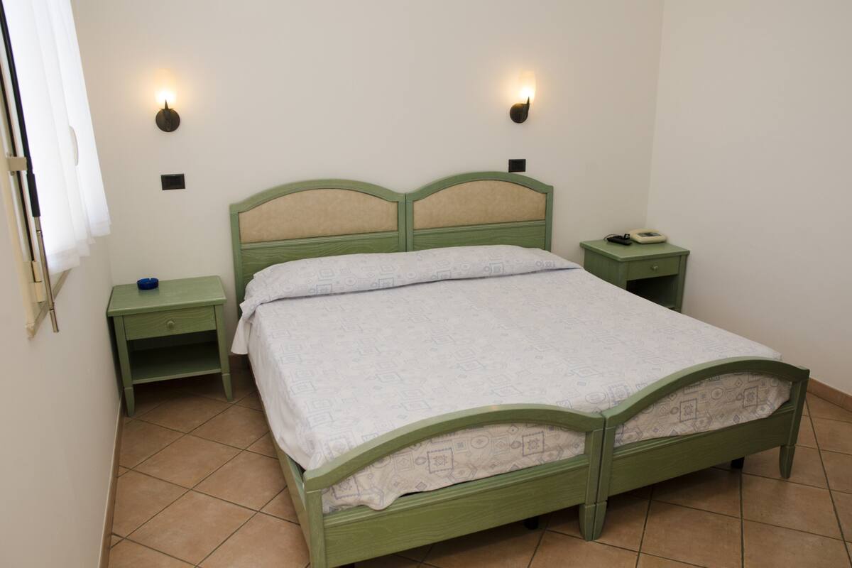 Uma cama de casal verde com colcha branca, duas mesas de cabeceira e luminárias fixadas na parede.