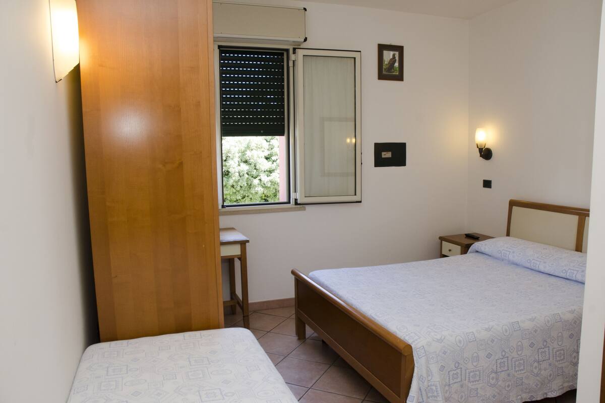 Quarto com duas camas, janela com persianas, mesa de cabeceira e luminária na parede.