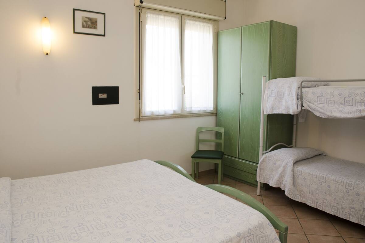 Quarto com beliches, guarda-roupa verde, uma cadeira e janela com cortinas.