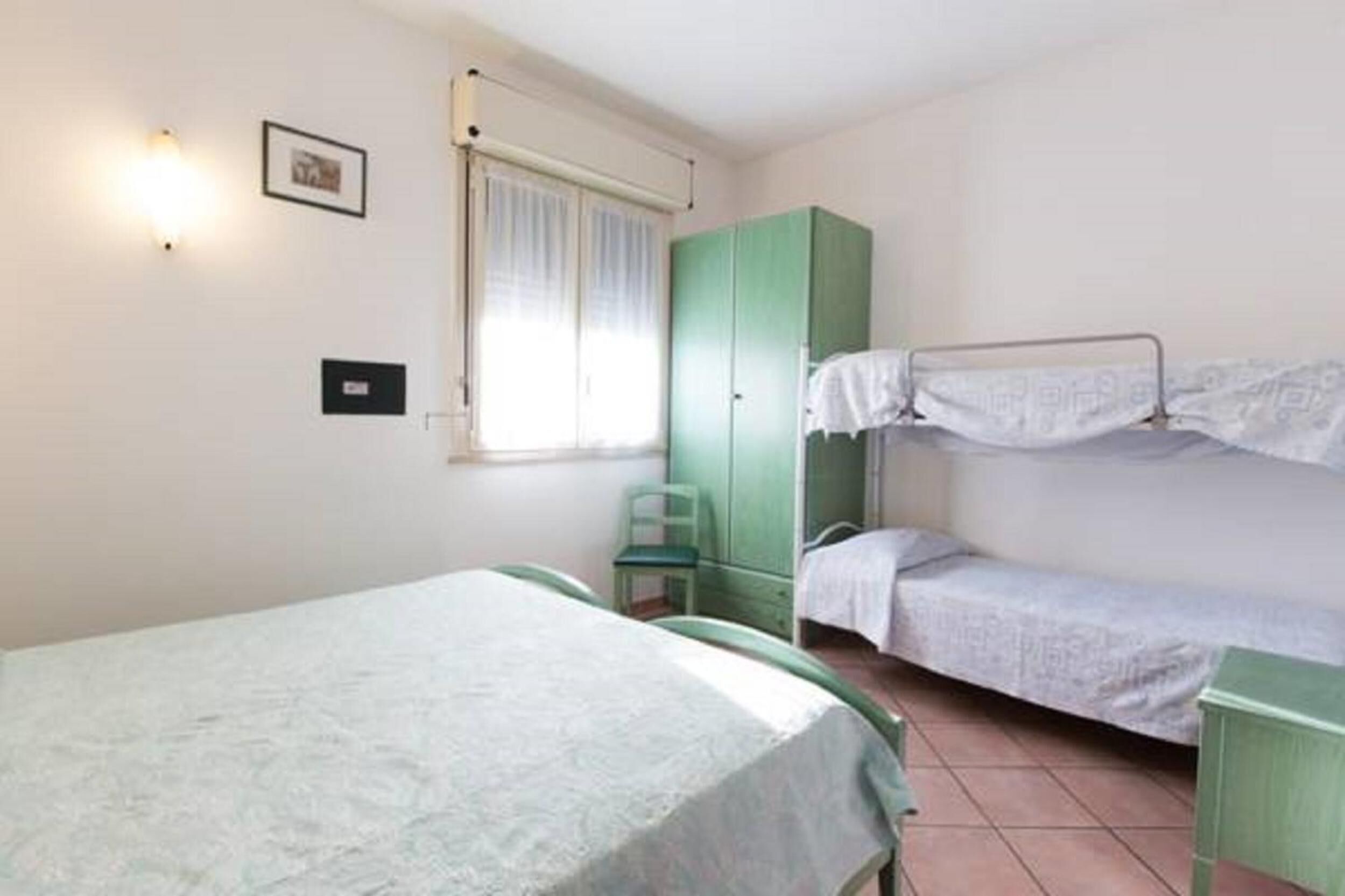 Une chambre avec un lit superposé, un lit simple, une armoire verte, une chaise et une fenêtre avec des rideaux.