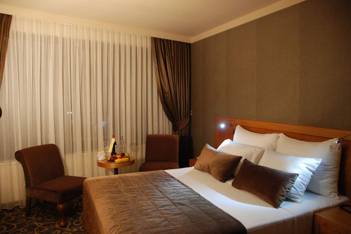 A hotel room with a large bed, two armchairs, a small table with a bottle and fruit, and a view through sheer curtains.
