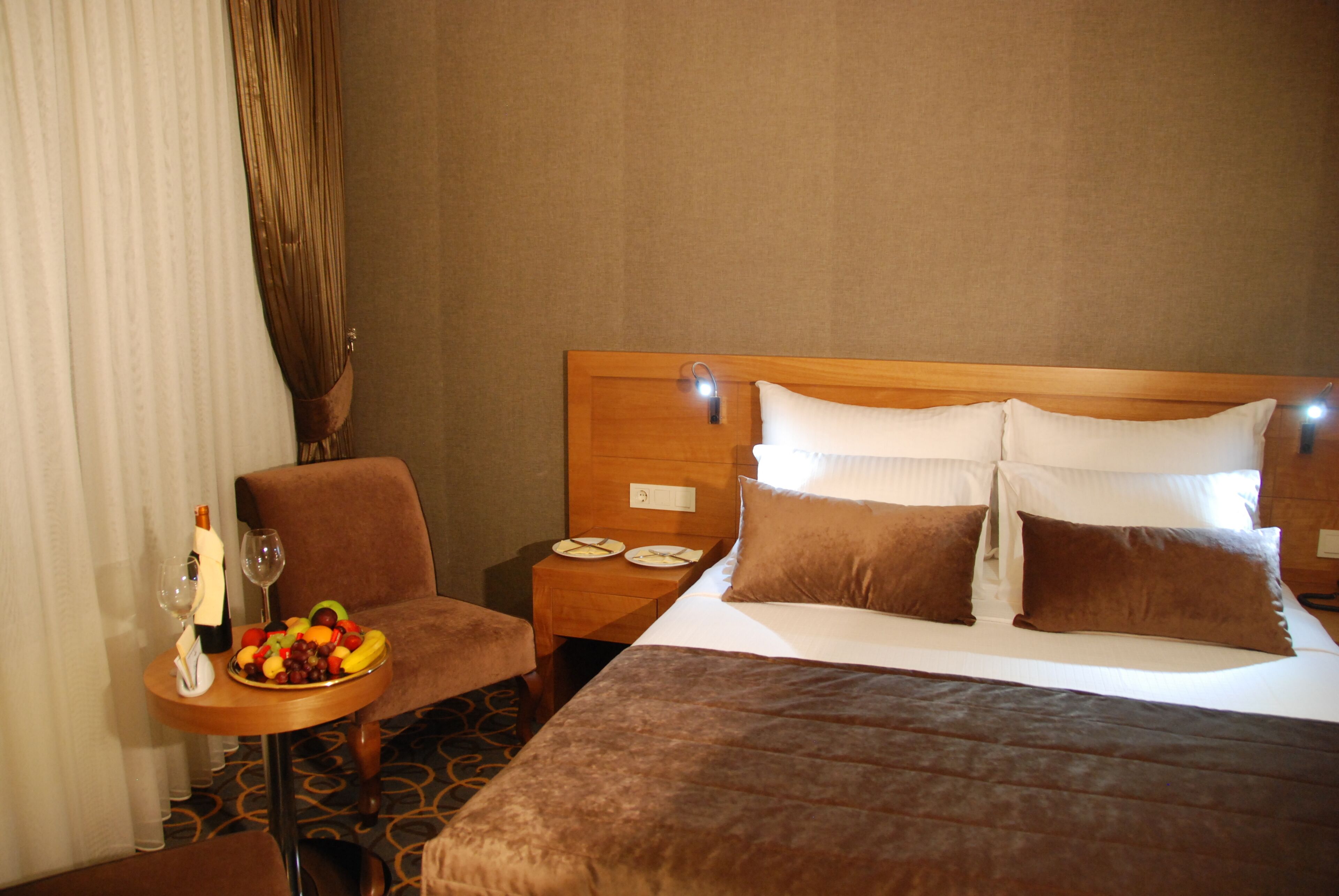 Habitación de hotel con una cama grande, una mesita de noche de madera con un cuenco de frutas, una botella de champán y una copa de vino.