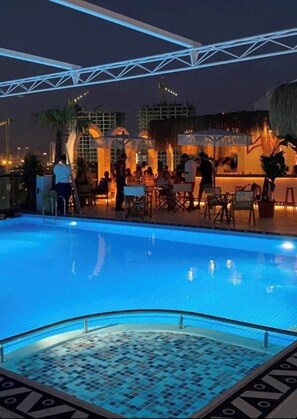 Zona de piscina al aire libre con solárium empinado, rodeada de sillones y sombrillas, y vistas del horizonte urbano nocturno.