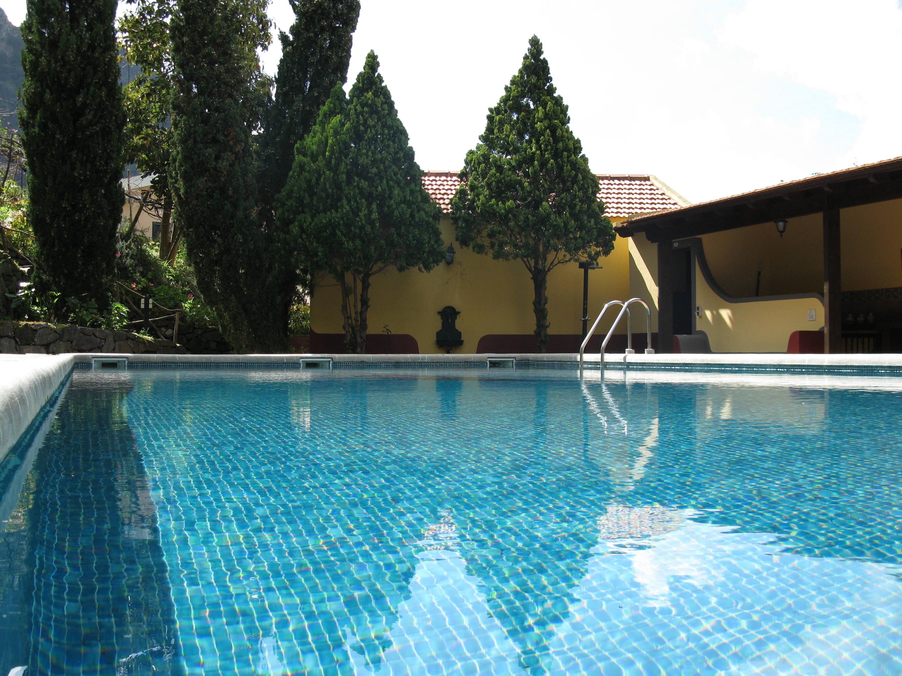Une piscine à l’eau d’un bleu cristallin, entourée d’arbres et bordée d’un bâtiment dont le toit est recouvert de tuiles.