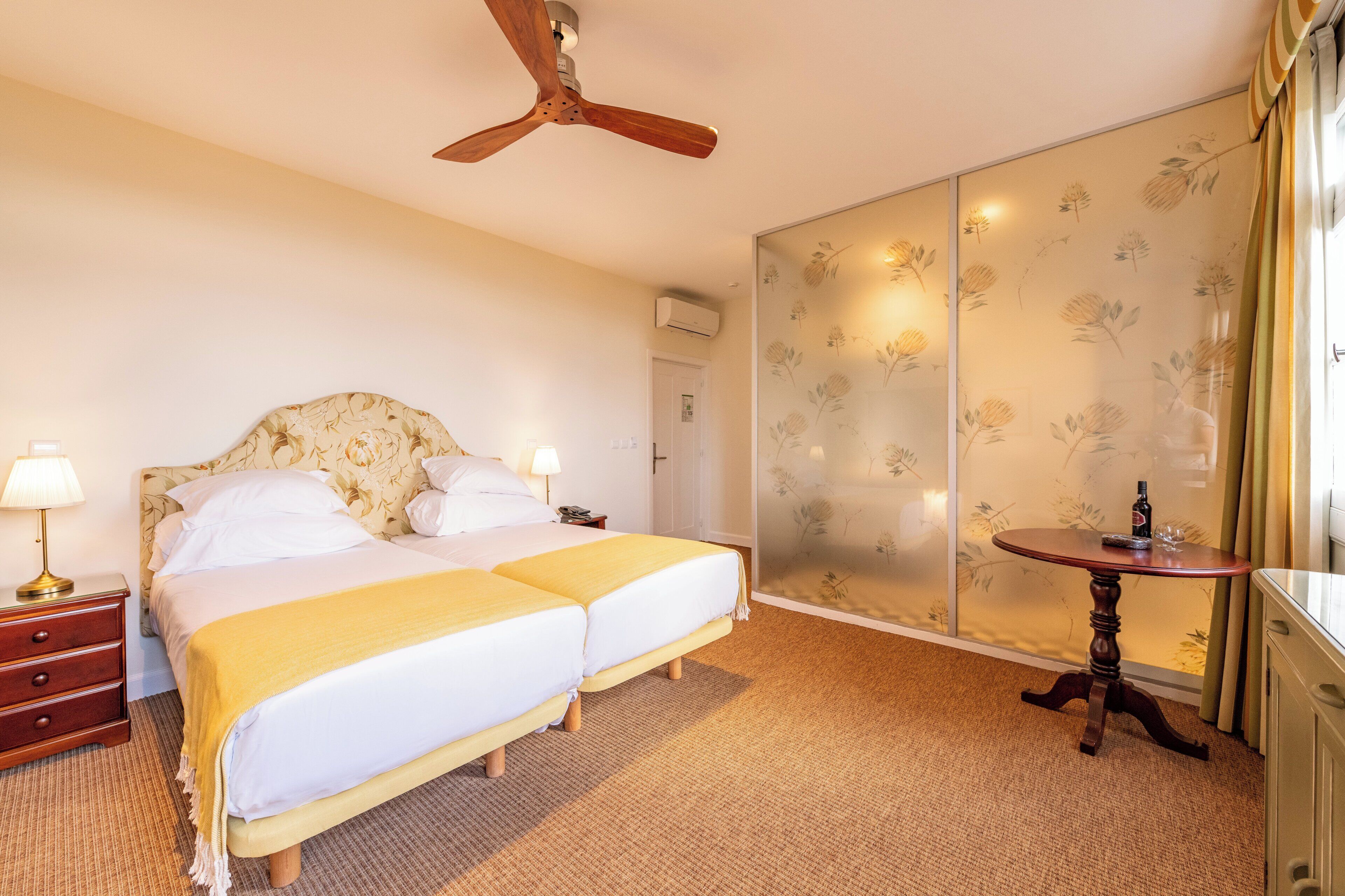 Ein Hotelzimmer mit zwei Betten, einem Deckenventilator, einem Tisch mit einer Flasche und einer Schiebetür mit floralem Wandpapier.