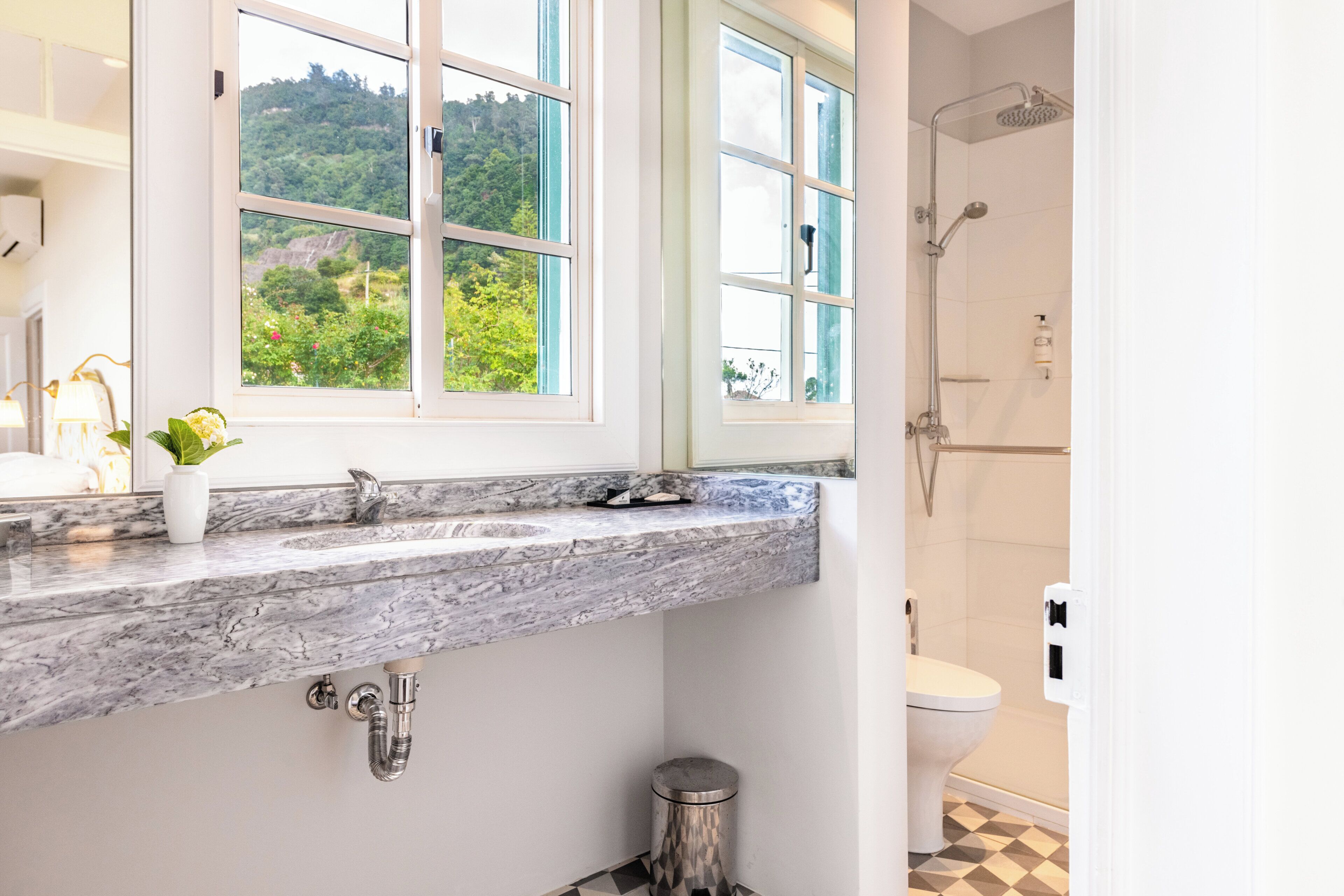A bathroom with a marble countertop, a window with a view of greenery, a shower, and a toilet.