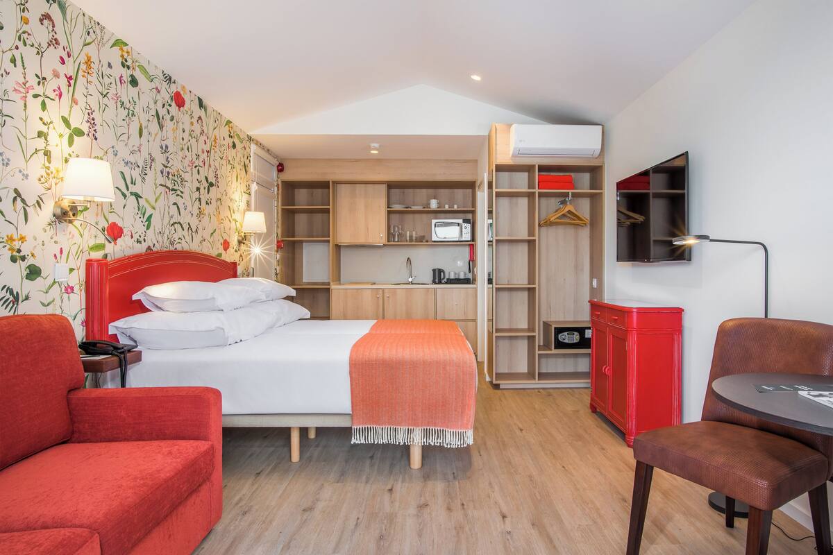 A hotel room with a bed, a red armchair, a wooden desk, a red cabinet, and a wall-mounted TV.