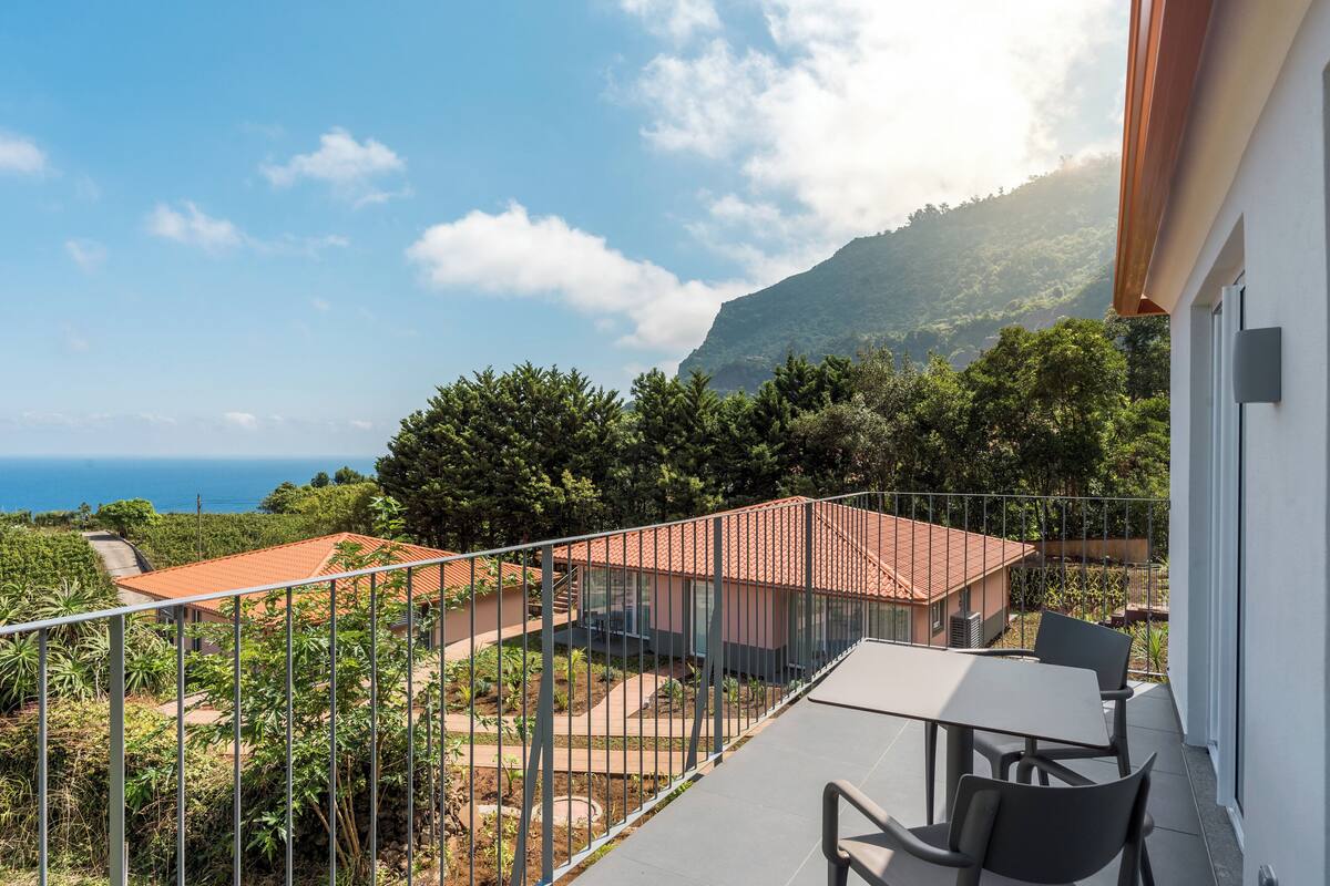 A balcony with a table and chairs, overlooking a mountainous landscape and a body of water.