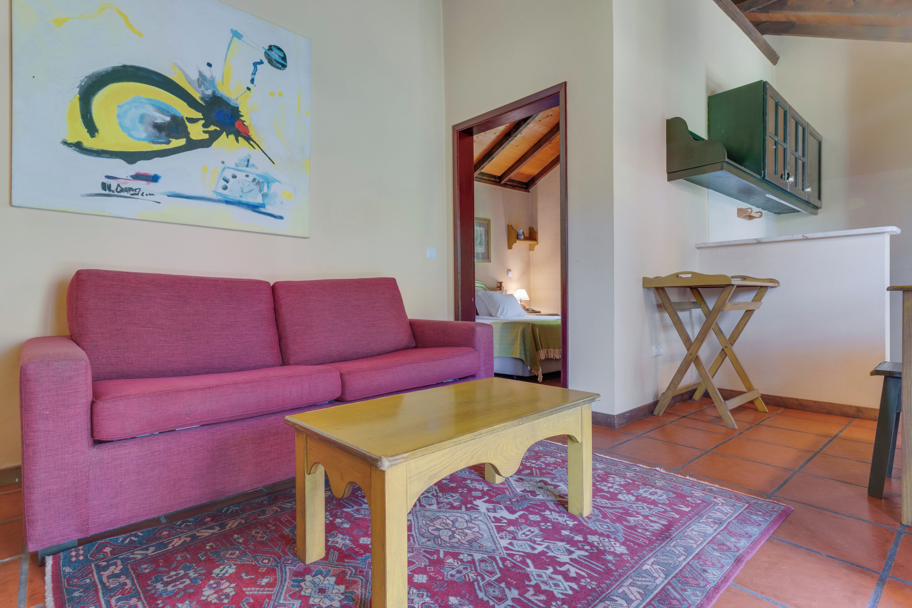 Una sala de estar con un sofá rojo, una mesa de centro de madera, un tapete estampado, un cuadro en la pared y una zona de cocina con un armario verde y una mesa plegable de madera.