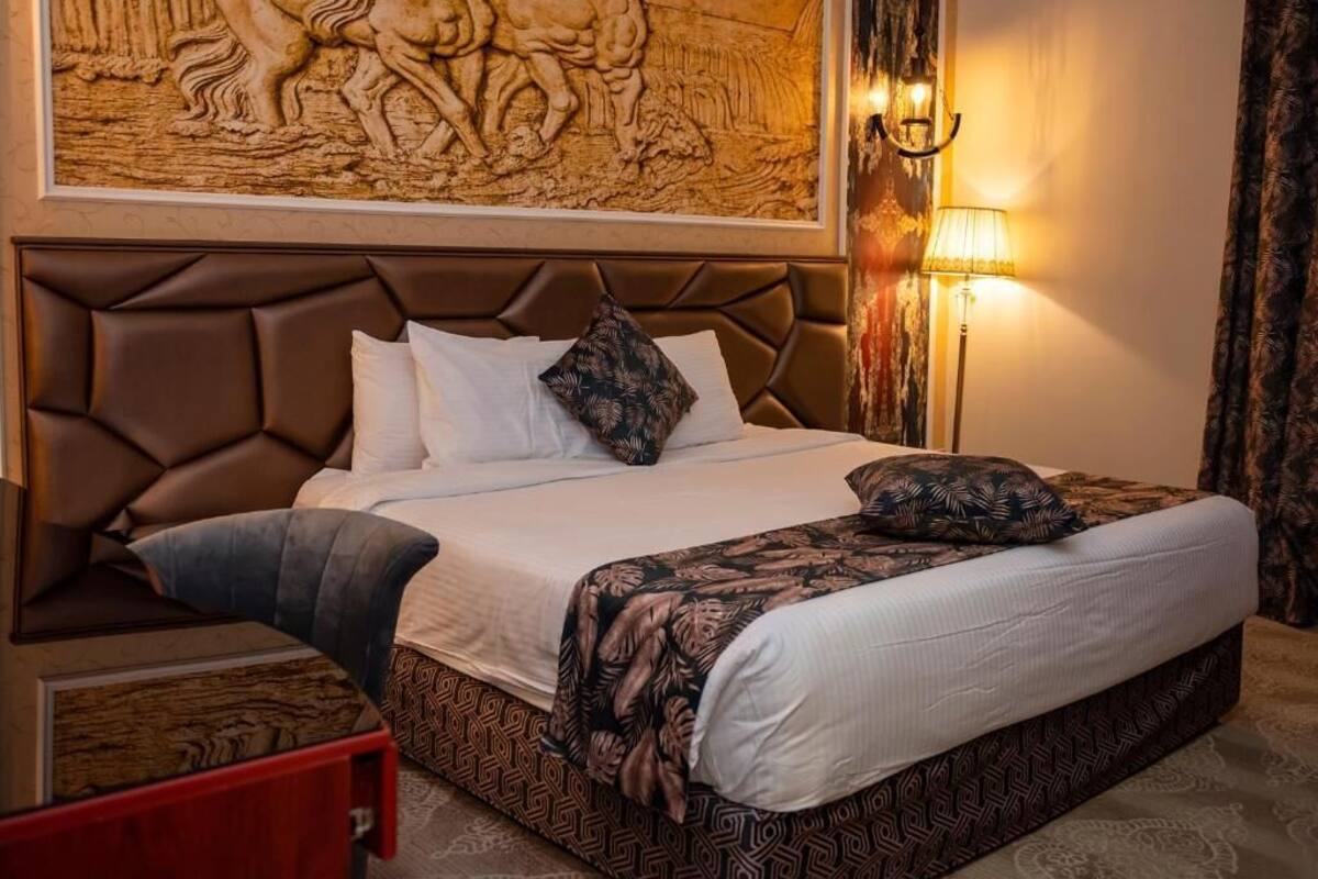 Una habitación de hotel con una cama grande, cabecero, lámpara de pared y una mesita de noche roja.