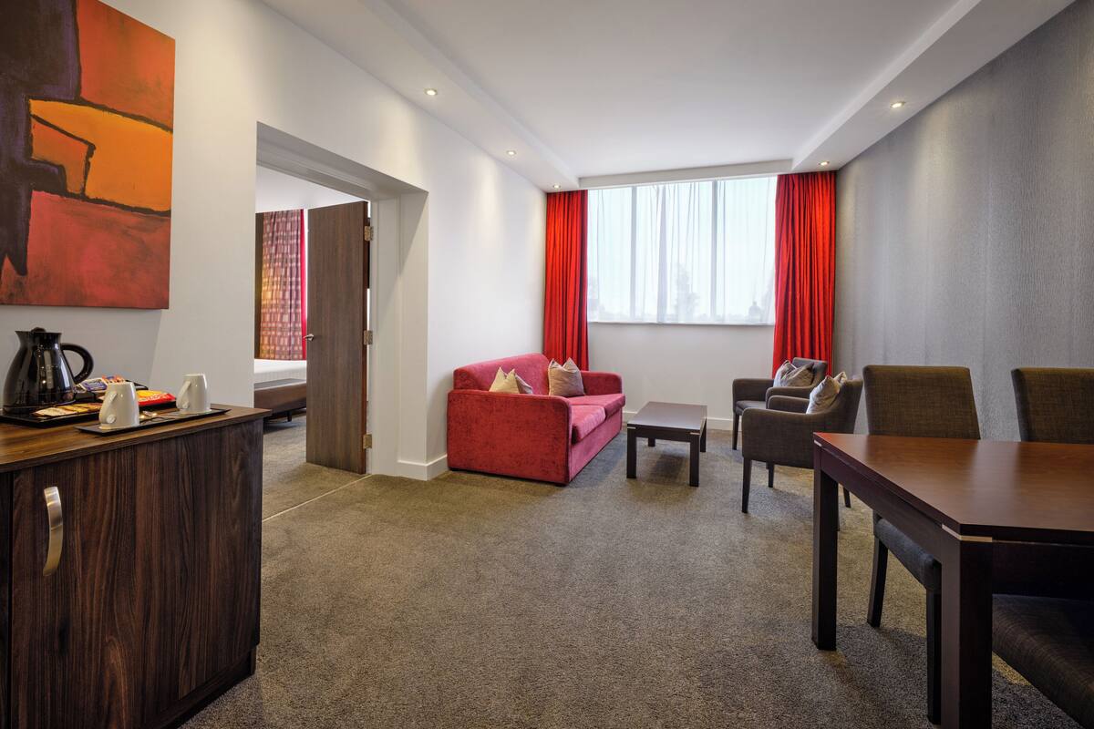 A modern hotel room with a wooden desk, a red sofa, a dining table, and a large window with red curtains.