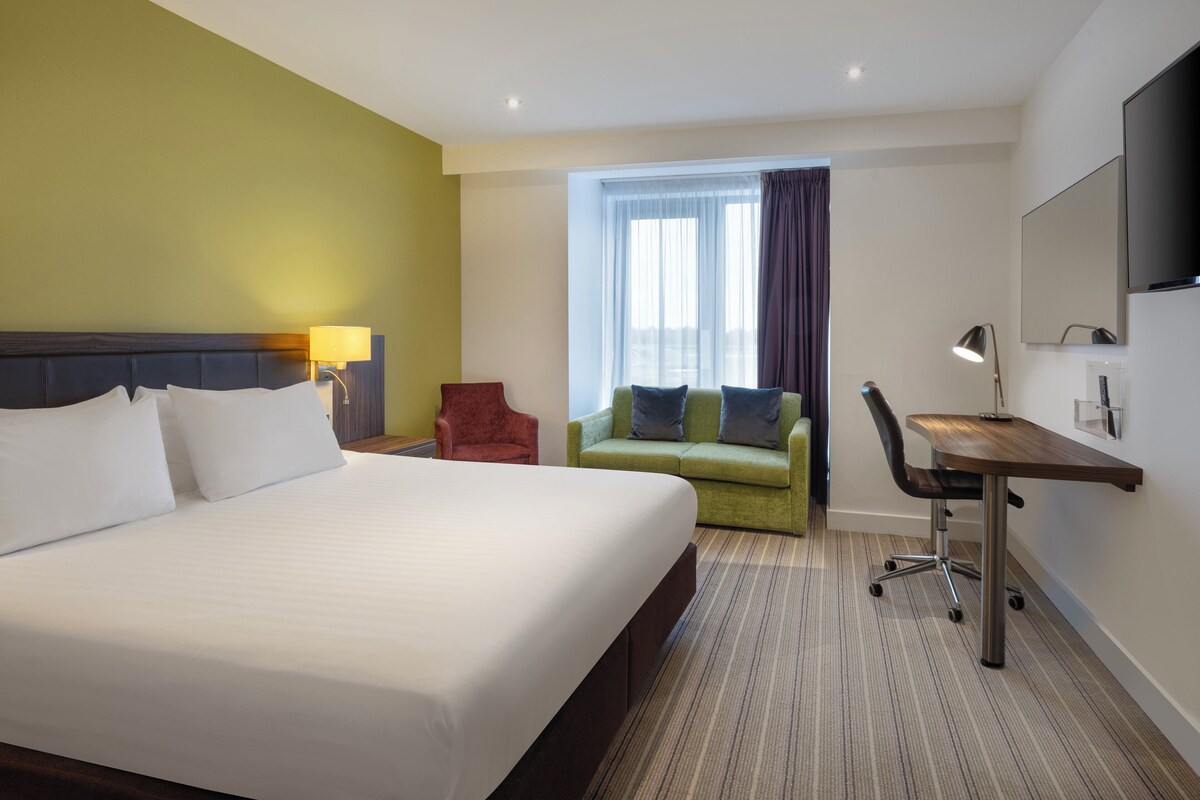 A hotel room with a large bed, a green sofa, a desk with a chair, and a TV mounted on the wall.