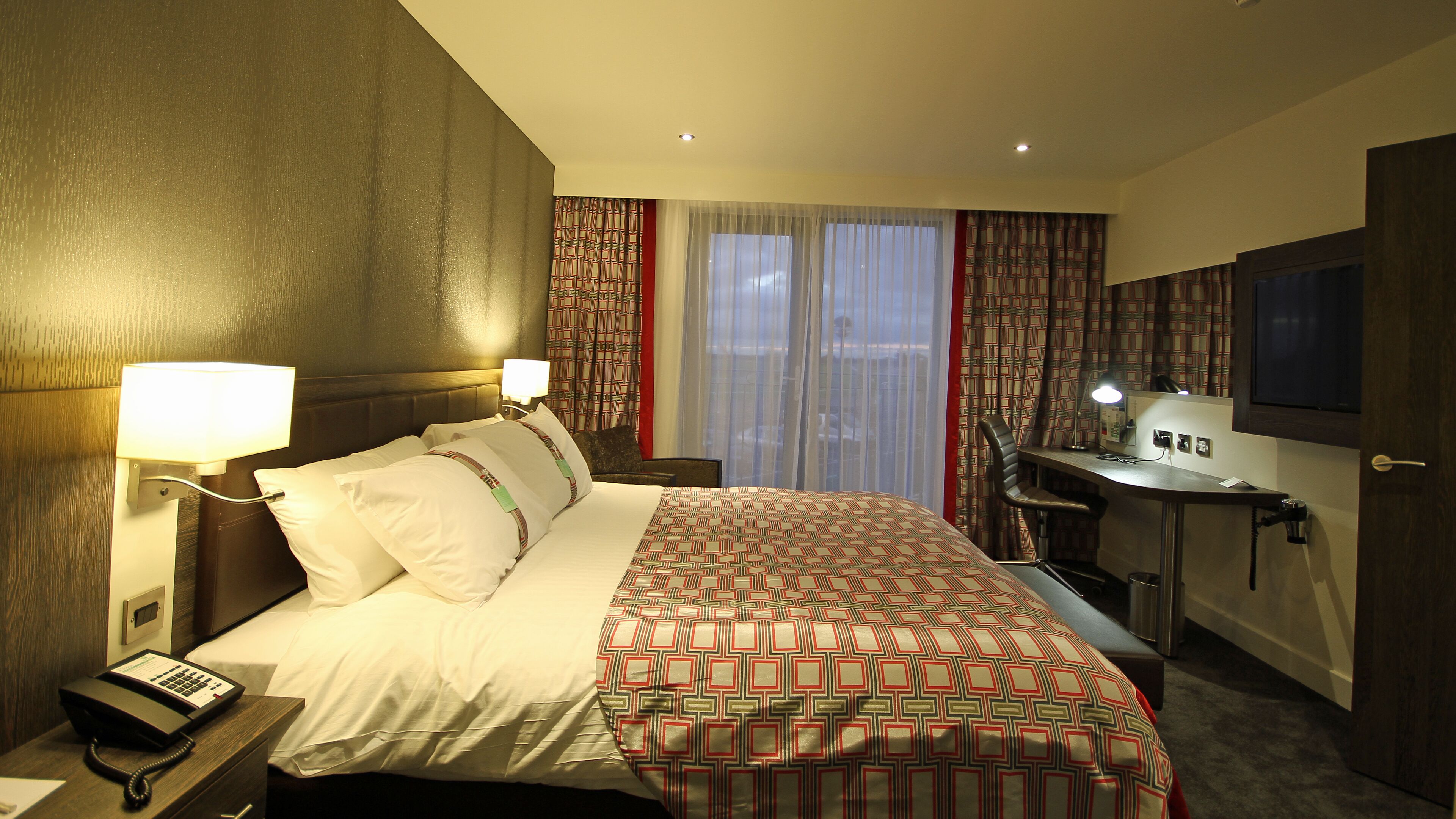 A hotel room with a bed, bedside lamps, a desk with a computer, a telephone, and a television.