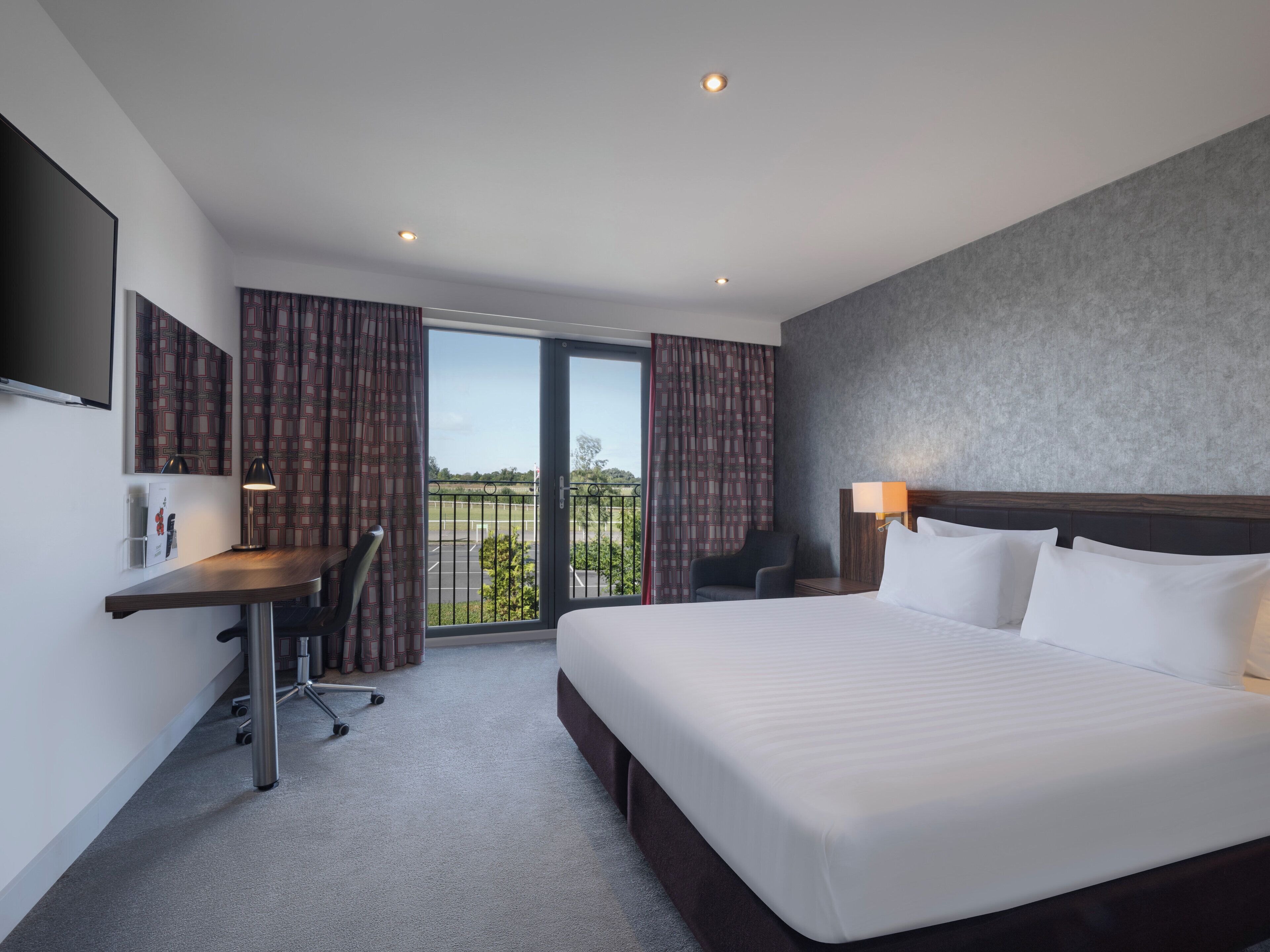 A hotel room with a large bed, a desk with a chair, a TV mounted on the wall, and a view of the outdoors through the window.