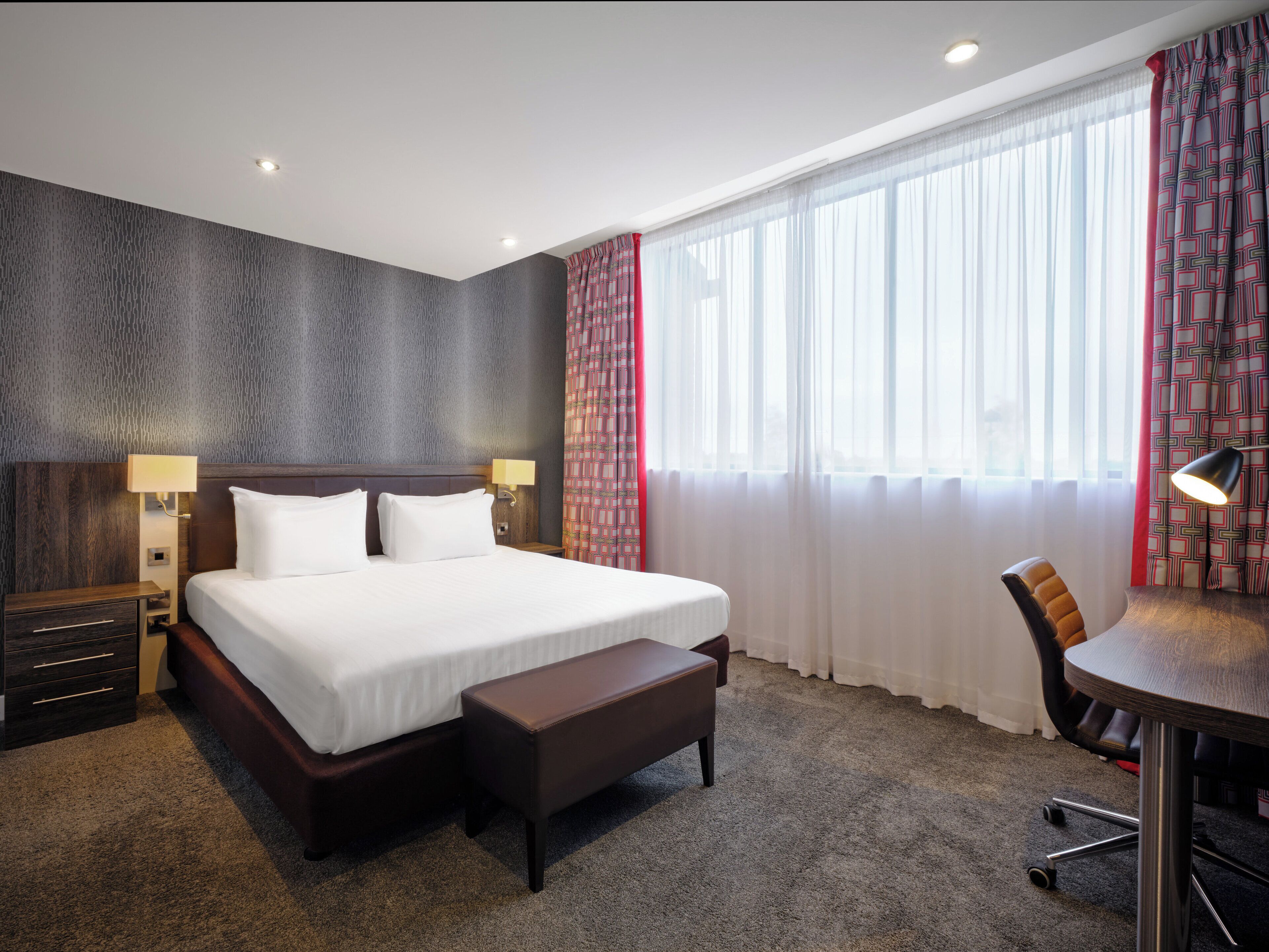 A hotel room with a large bed, two bedside tables with lamps, a desk with a chair, and a large window with sheer curtains.