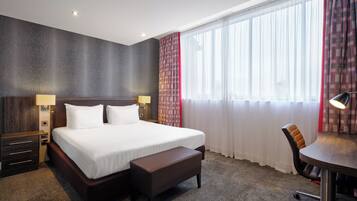 A hotel room with a large bed, two bedside tables with lamps, a desk with a chair, and a large window with sheer curtains.