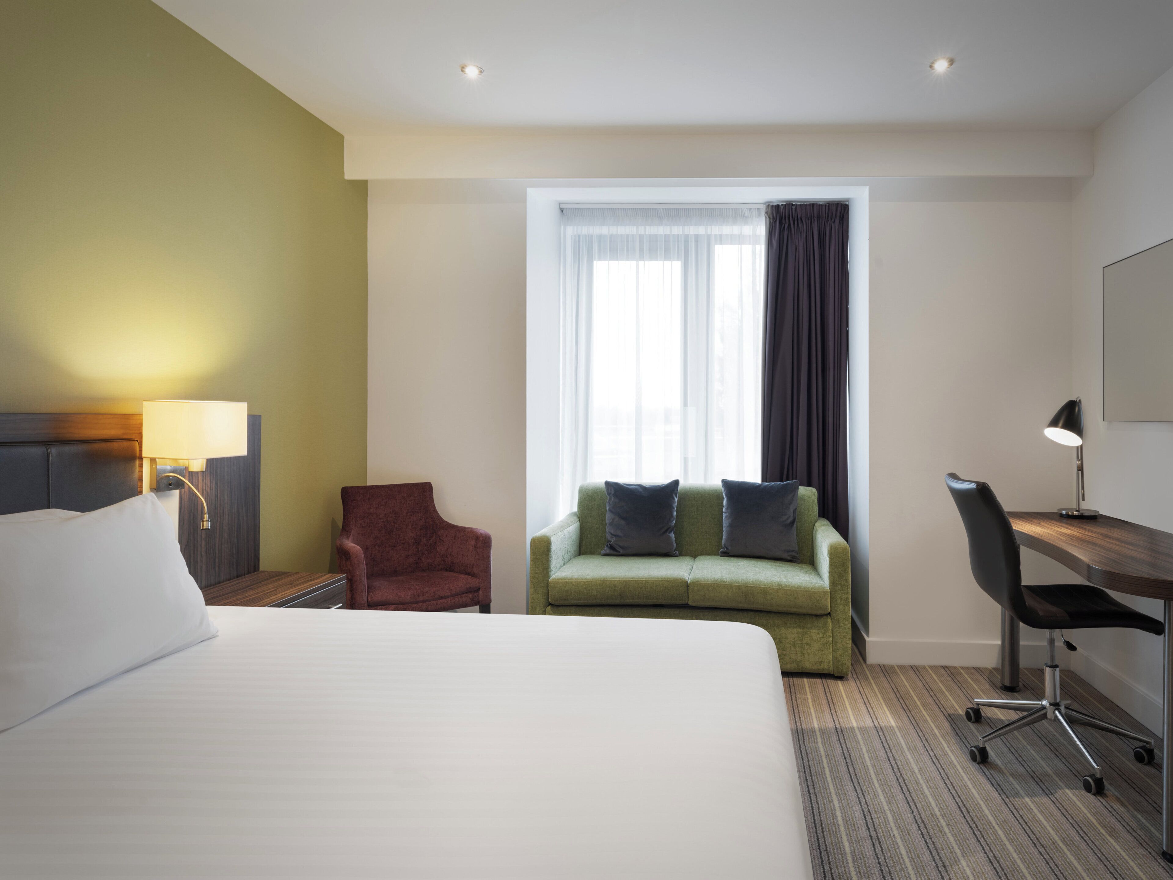 A hotel room with a large bed, a green sofa with two pillows, a red armchair, a desk with a chair, and a window with curtains.