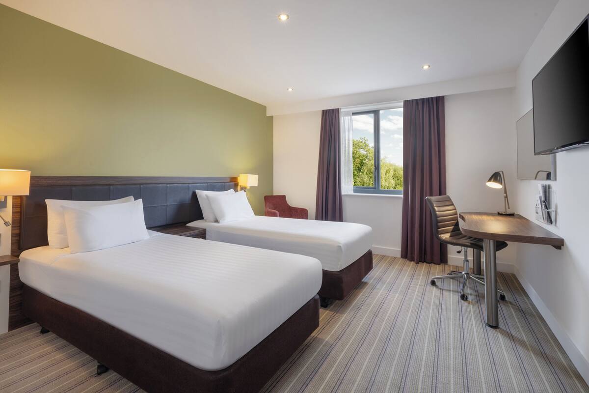 A hotel room with two beds, a desk, a chair, a TV, and a window with curtains.