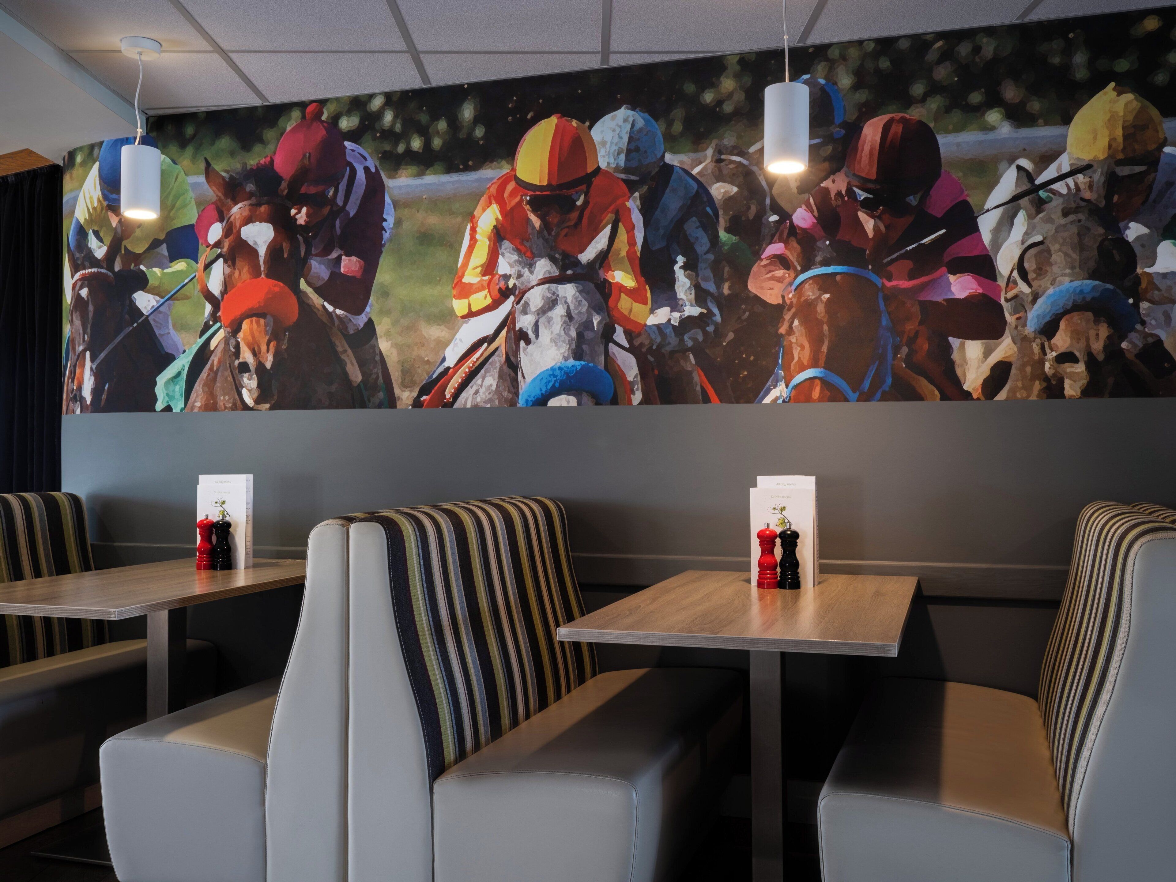 A restaurant with striped booths, tables with salt and pepper shakers, and a large horse racing mural on the wall.