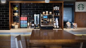 Interior moderno de una cafetería con una mesa de madera, sillas, una máquina de café, estantes con tazas de café y una pantalla de menú.