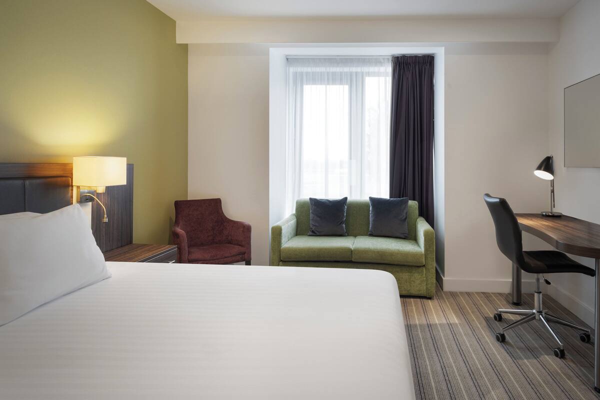 A hotel room with a large bed, a green sofa with two pillows, a red armchair, a desk with a chair, and a window with curtains.
